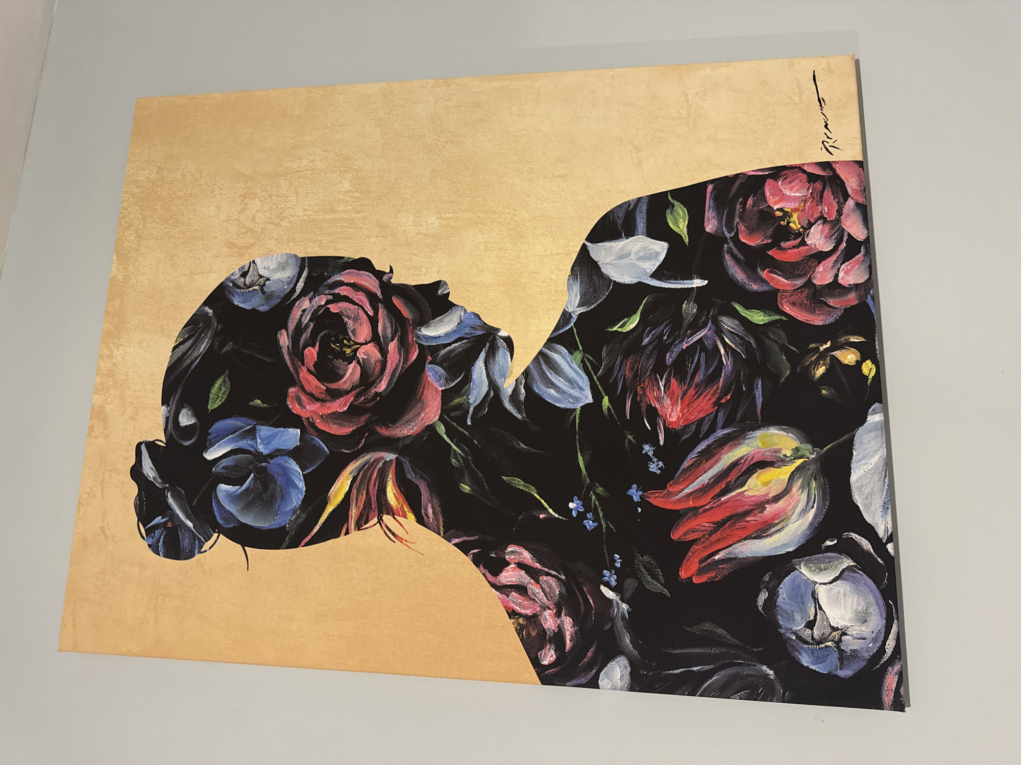 A painting hangs on a gray wall. The painting features a gold background. A silhouette of a person's head and neck is created by a dense arrangement of colorful flowers, predominantly roses and tulips, set against a black background. The signature of the artist is visible in the top right corner of the painting. The location is Unknown, Unknown.