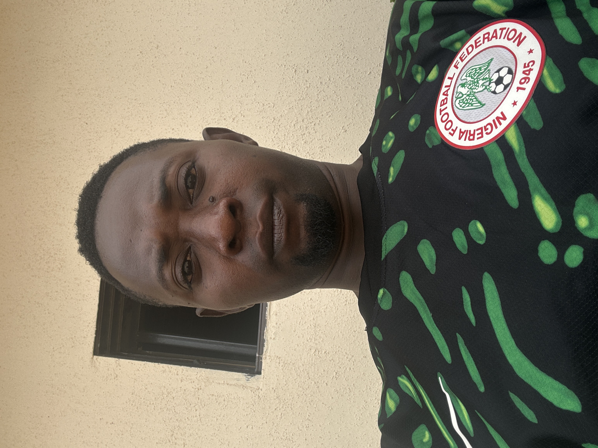 A medium shot shows a Black man in his late 30s facing the camera, centered in the frame. He has short, dark hair and a short beard. He is wearing a black t-shirt with a green and white abstract pattern and a circular crest with "Nigeria Football Federation 1945" on the left chest. Behind him is a textured, light-colored wall with a small, dark-framed window visible on the upper left. The lighting is natural and appears to be from an overhead source. The overall scene is a straightforward outdoor portrait.