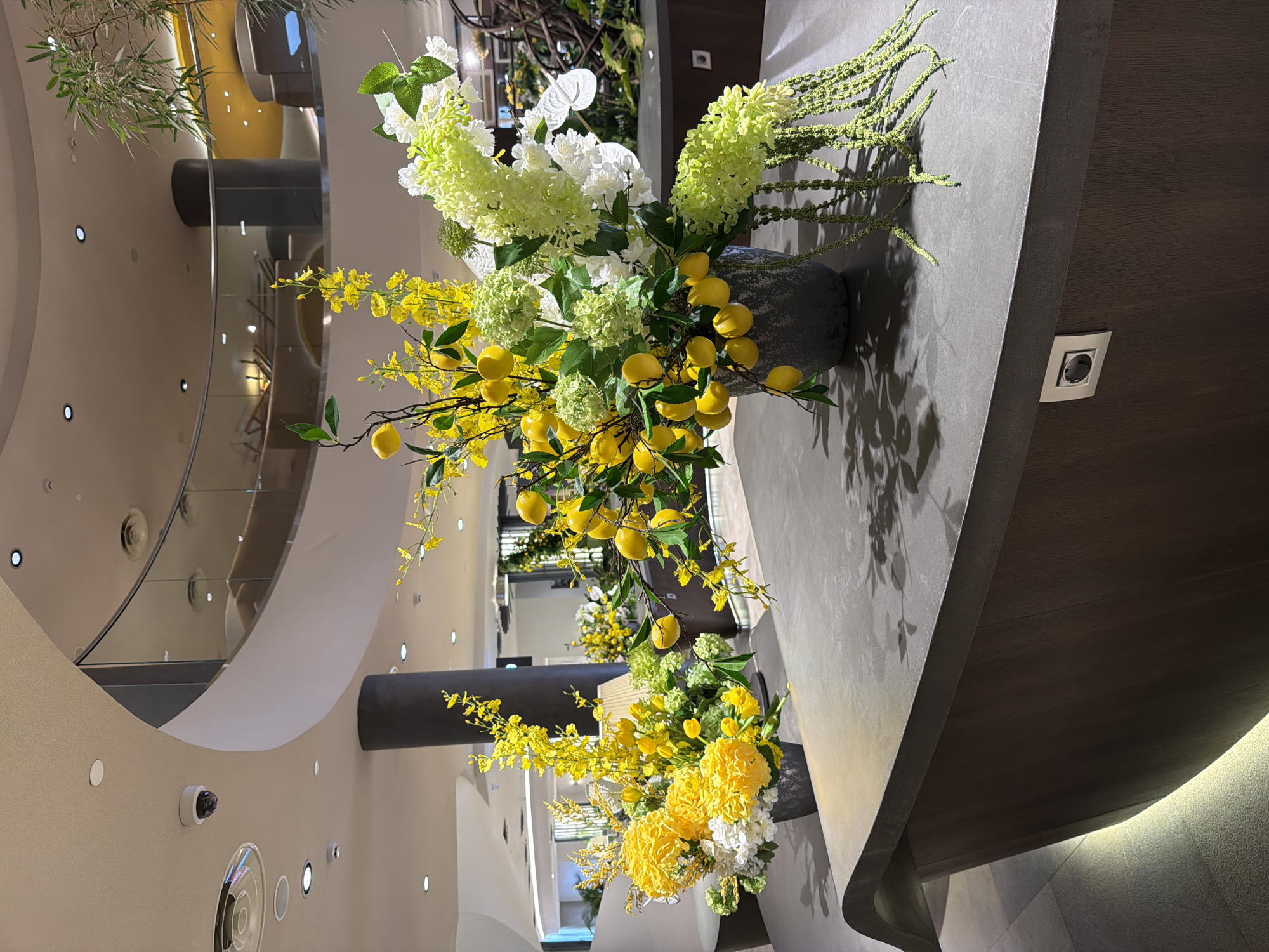 The image depicts floral arrangements in what appears to be a modern interior space.
Arrangements feature white and green hydrangeas, yellow flowers, and branches with yellow lemons. The arrangements are displayed in dark gray vases. One is prominently displayed at the corner of a countertop, the second is positioned further back. The lighting in the area casts shadows of the flower arrangements on the countertop.
In the background is a multi-level architectural space with a curved design, possibly a shopping mall or large building interior. There are lights, cameras and other equipment visible on the walls and ceiling. It is not possible to determine the specific location in Moscow, Russia from the image.
