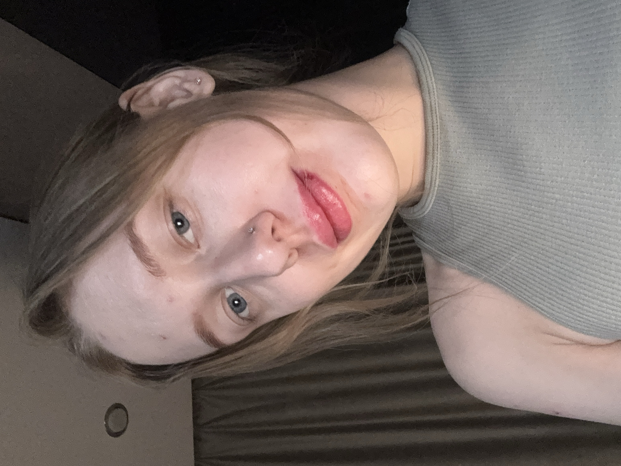 A young woman with light blonde hair and fair skin is captured in a close-up, tilted selfie. Her light-colored eyes are open, and she has piercings in her nose and ear. She is wearing a light green ribbed tank top. The background is partially dark, with hints of a wall, door, and textured fabric. There is no evidence to suggest the location is Rostov-on-Don, Russia.