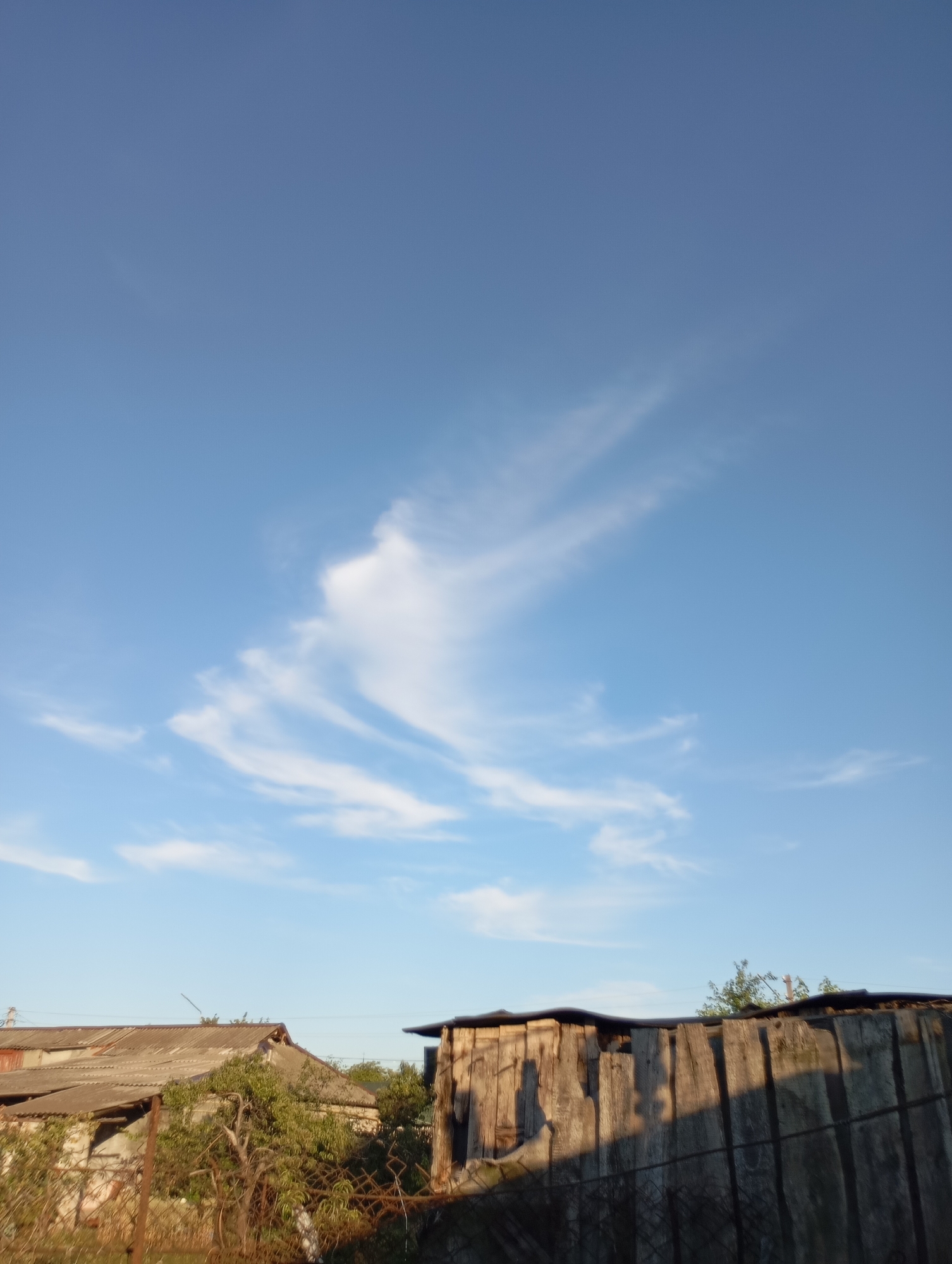 The image depicts an outdoor scene with a clear blue sky and scattered wispy clouds. In the foreground, there are two dilapidated structures. The structure on the left appears to be a house with a corrugated metal roof and weathered walls. The structure on the right is a wooden fence or shed made of vertical planks, showing signs of age and decay, with shadows cast across its surface. Vegetation, including trees and bushes, is visible between and around the structures. A rusty metal fence runs along the bottom of the image. The lighting suggests it is either early morning or late afternoon, with sunlight casting long shadows. The overall impression is of a rural or neglected urban environment.