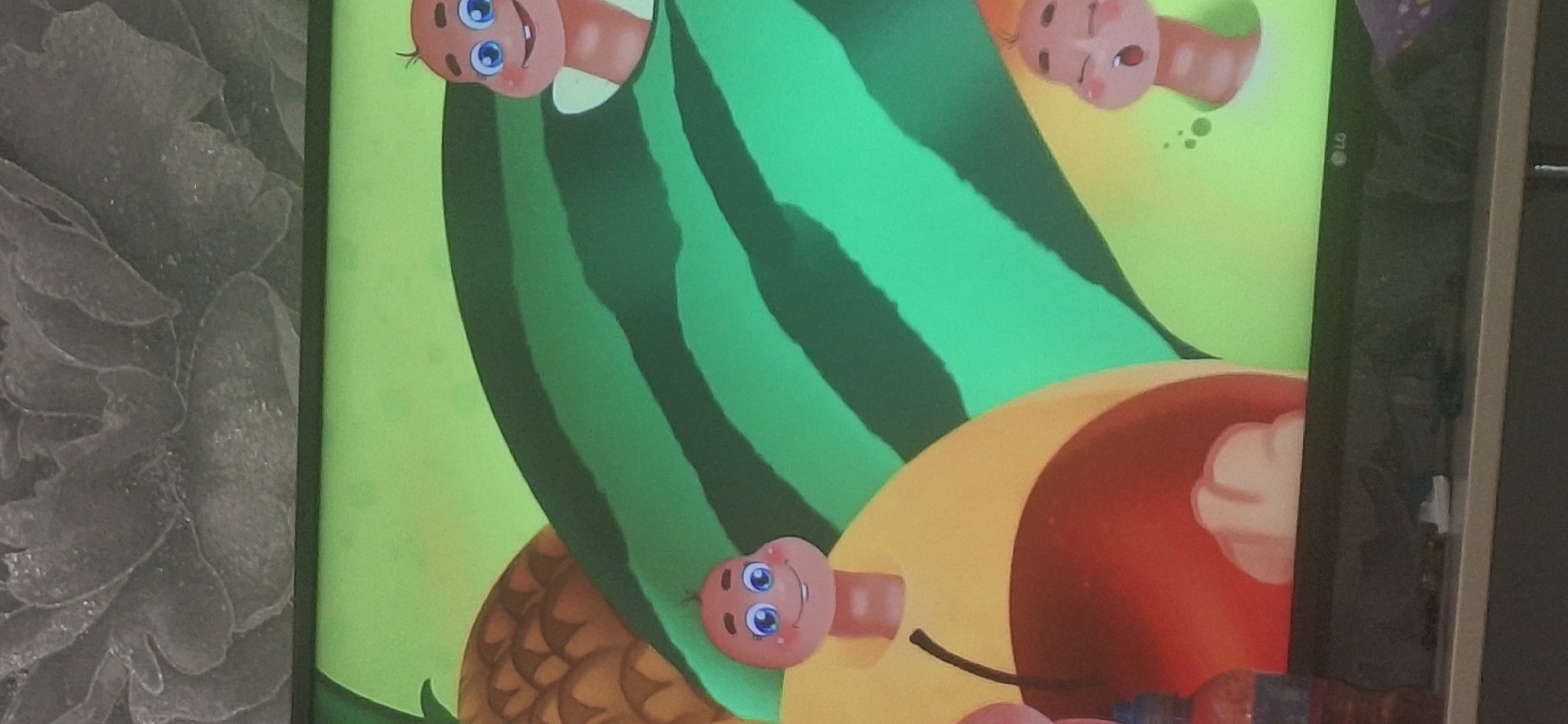 The image is a photograph of a television screen displaying a cartoon animation. The animation features several cartoon caterpillars with human-like faces, some peeking out from behind various fruits. In the foreground, on the left side of the screen, a portion of a yellow and red apple is visible, with a caterpillar head emerging from it. Behind the apple, part of a pineapple can be seen. A large watermelon with green stripes dominates the center and right side of the screen, with another caterpillar head emerging from it. A third caterpillar head, with an open mouth, is visible near the bottom right corner, emerging from what appears to be a light green background. The background of the animation is a bright, solid green. The television is placed on a dark surface, possibly a stand or shelf. The wall behind the television has a textured, grey pattern. The presence of a cat toy on the shelf suggests a domestic setting. There is no information in the image to indicate a location in Bila Tserkva, Ukraine.