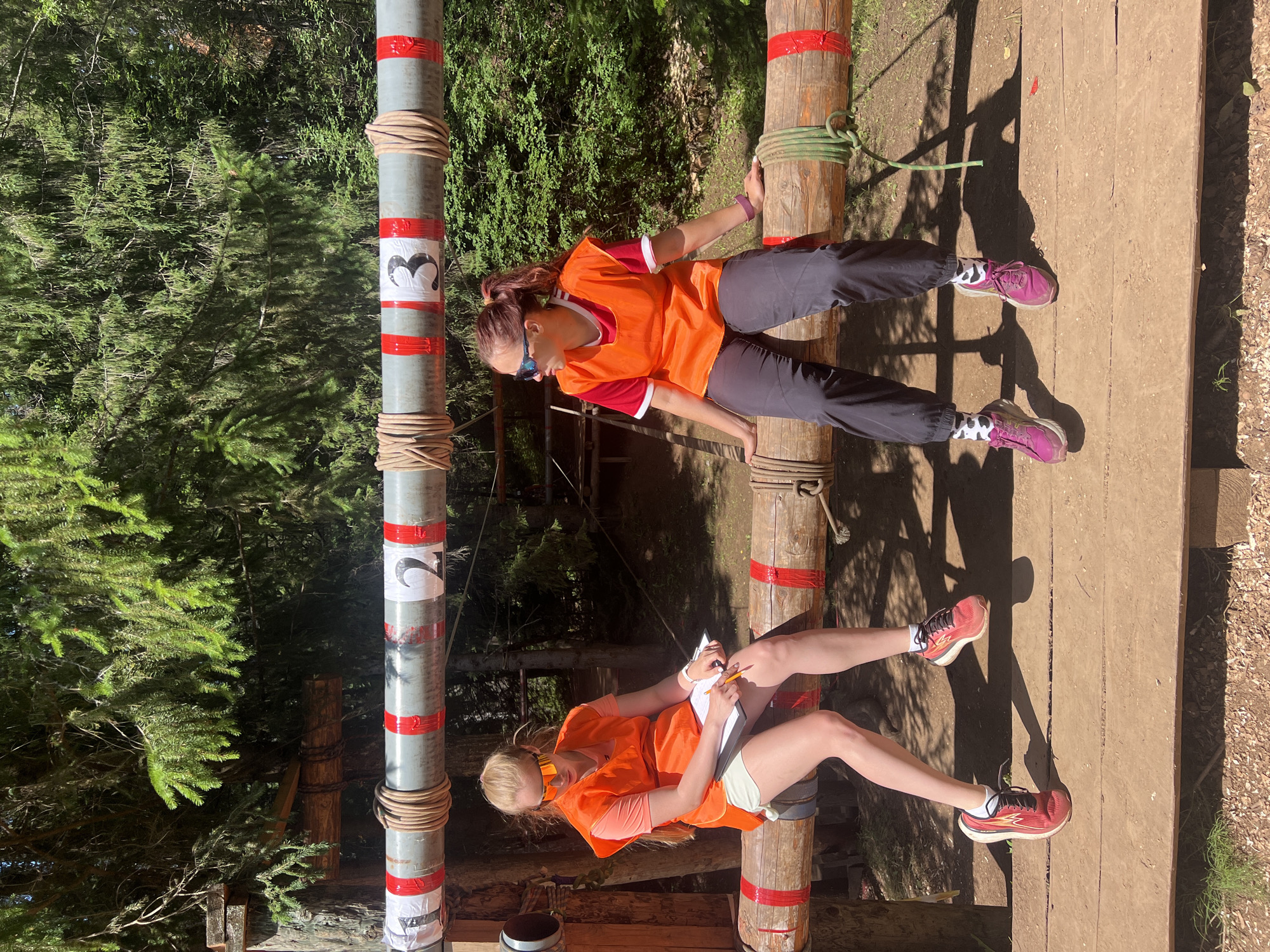 Two young women are participating in an outdoor obstacle course. Both are wearing orange vests over athletic tops. The woman on the left has her arms extended, holding onto vertical poles wrapped in rope and marked with red tape and white circles containing black numbers "3" and "2". She wears sunglasses and dark pants. The woman on the right is sitting and writing on a clipboard with a pencil. She wears orange sunglasses and light-colored shorts over leggings. Her legs are extended, resting on a wooden platform. Her feet are adorned with socks featuring a black and white cow-print pattern, and she wears pink athletic shoes. The scene is set in a forest with trees and dappled sunlight, suggesting a sunny day. The obstacles appear to be part of an adventure park or training facility.