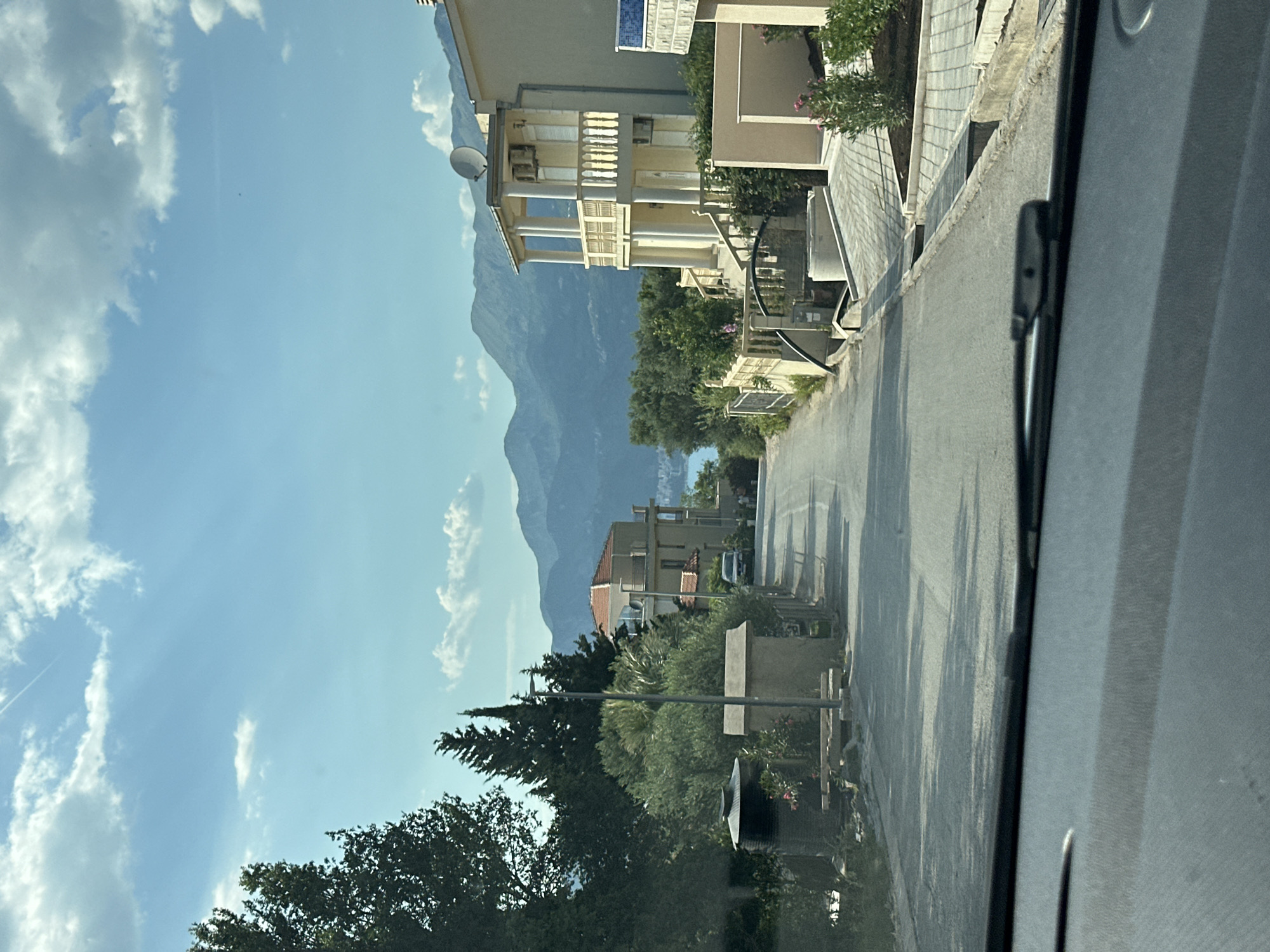 This media file captures a view from inside a vehicle, looking out onto a street lined with buildings and lush greenery. The scene is dominated by a large, imposing mountain range in the background, its peaks shrouded in a light haze. The sky above is a vibrant blue, dotted with scattered white clouds, suggesting a clear and pleasant day.
On the right side of the frame, a paved road stretches into the distance, casting long shadows from unseen trees, indicating it is likely late afternoon or early morning. Adjacent to the road, there are several multi-story buildings, some with balconies and decorative elements, suggesting a residential or vacation area. Dense green foliage, including trees and bushes, flourishes between the buildings and along the roadside, adding a natural element to the urban landscape. A satellite dish is visible on one of the buildings.
There are no people clearly visible in the frame, and the focus is on the static elements of the environment. The overall impression is one of a quiet, sunny day in a picturesque location, possibly a coastal town given the presence of mountains and what appears to be distant water in the far background, though this is not clearly discernible. The media file is oriented vertically.