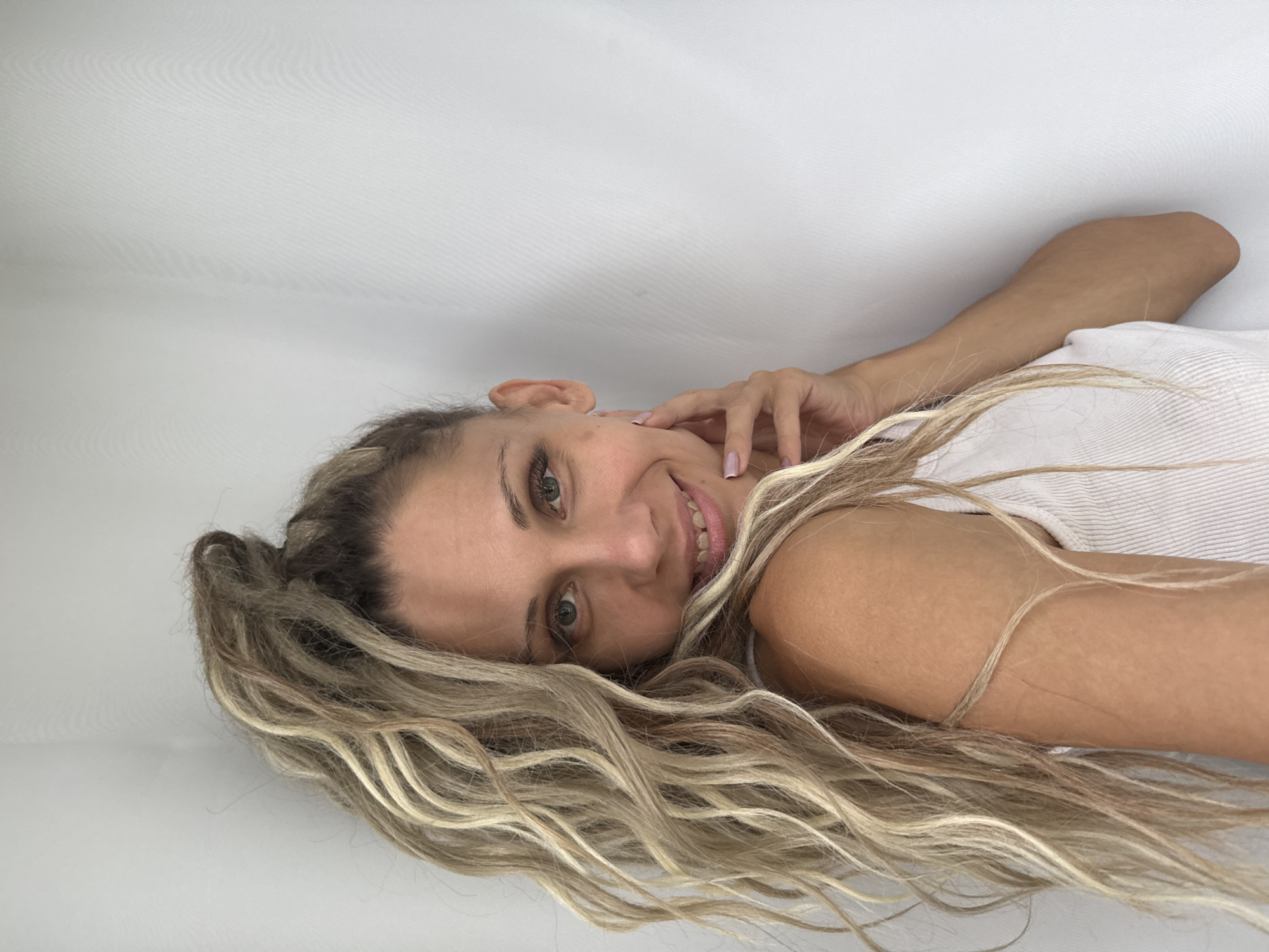 A medium shot captures a woman with blonde dreadlocks pulled into a high ponytail, against a white background. She is turned towards the viewer, with her left hand resting on her cheek and her right shoulder visible. Her hair is a mix of blonde and brown tones, with long, wavy strands cascading down her left side. She wears a white ribbed tank top. Her eyes are green, and she has a slight smile, revealing some of her teeth. The lighting is bright, creating a soft glow on her skin. The environment appears to be indoors, with a plain white curtain or wall serving as the backdrop. The location in the city of Rusanovo, Russia, is not evident from the visual information provided in the image.