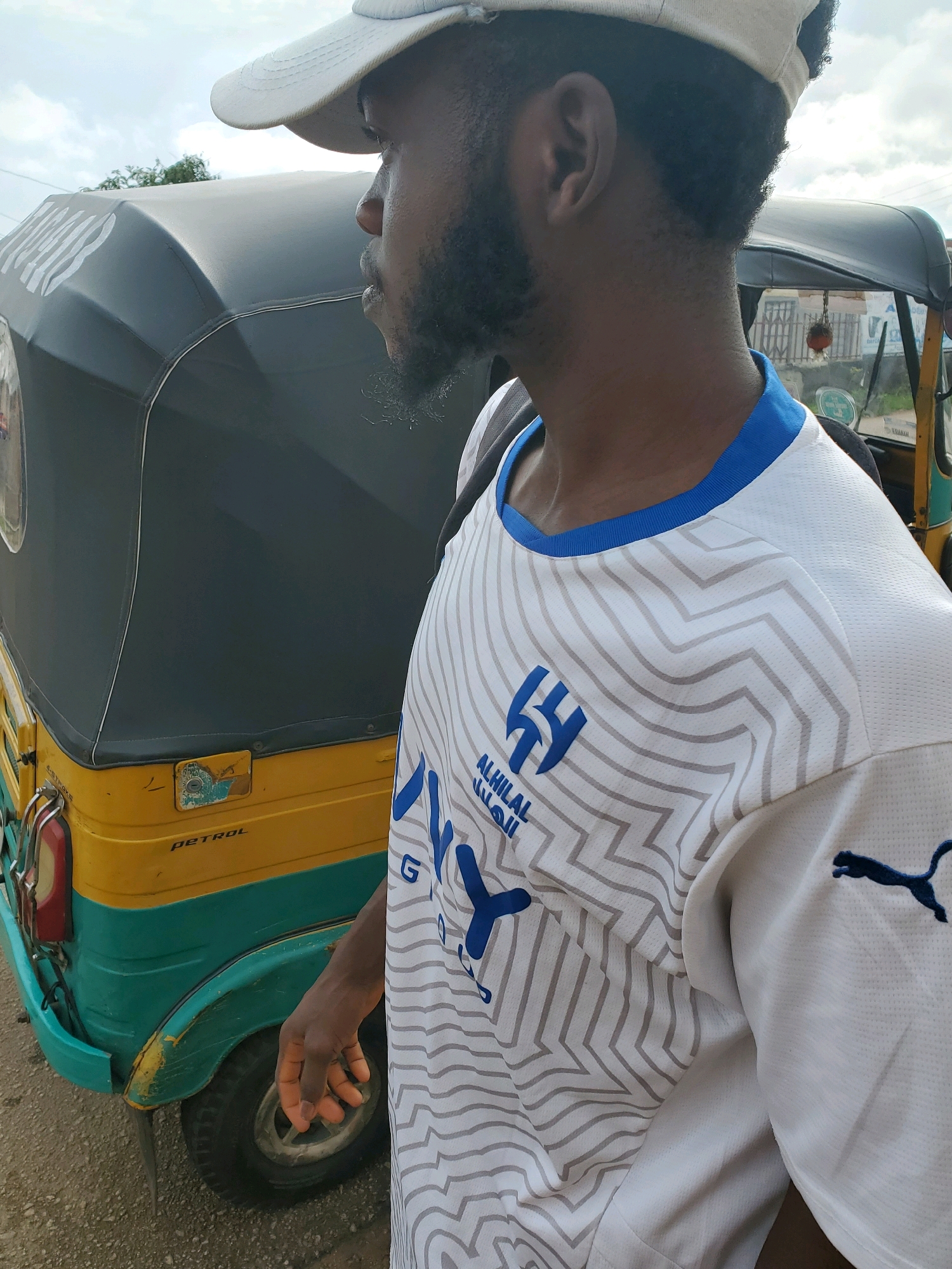 In Jos, Nigeria, a man wearing a white and grey patterned athletic shirt with blue trim stands outdoors next to a yellow and teal auto-rickshaw. The man has dark skin, short black hair, and a beard, and is wearing a light-colored baseball cap. The auto-rickshaw has a dark grey canopy and the word "PETROL" is visible on its side. The scene appears to be on a paved surface, likely a street or road, under an overcast sky.