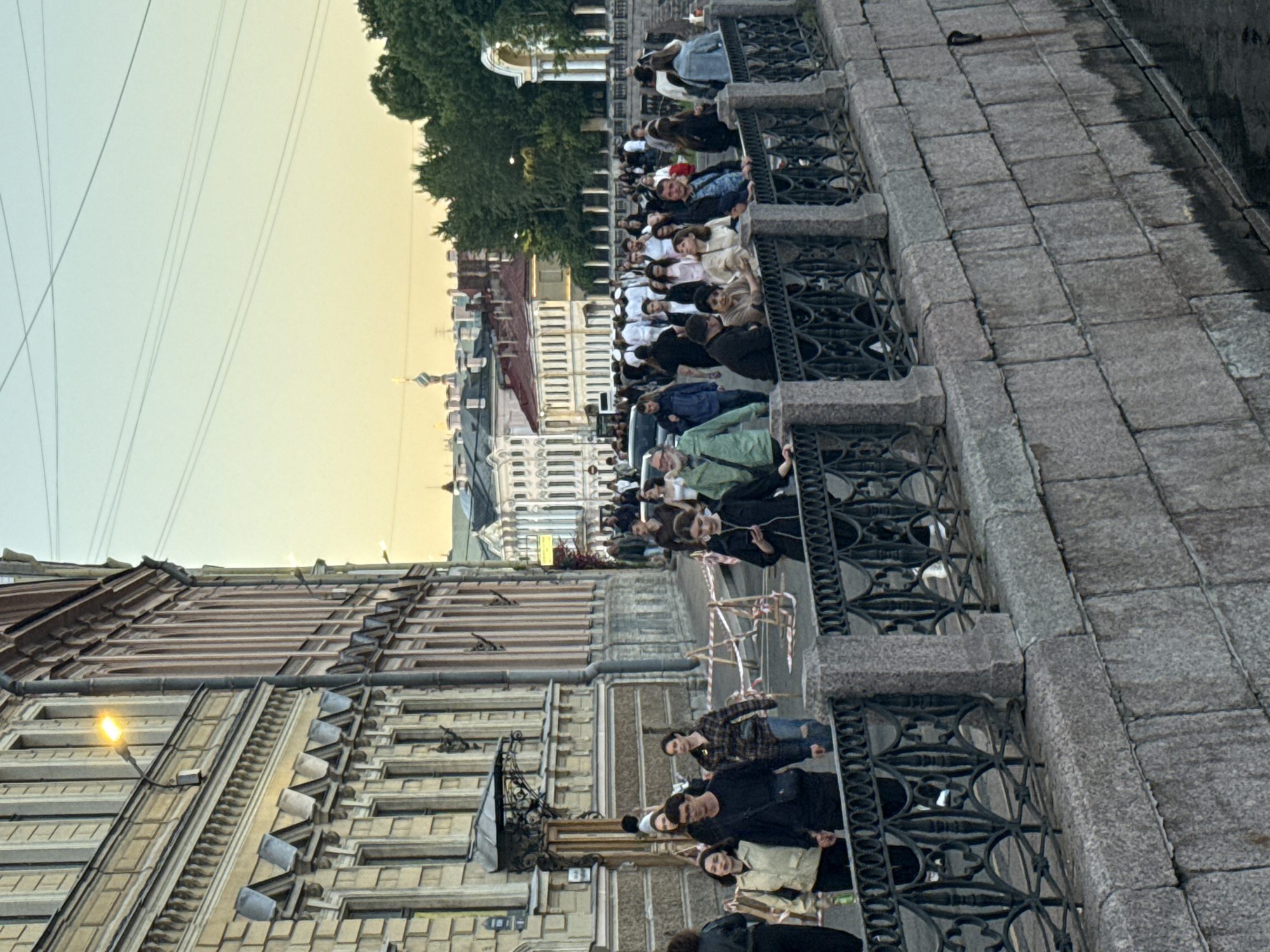 The image depicts a street scene along a canal in Saint Petersburg, Russia, during what appears to be the late afternoon or early evening, judging by the soft, warm light. The perspective is from a slightly elevated position, looking down towards a stone embankment and a canal.
On the left, a grand, multi-story building with classical architecture dominates the scene. Its facade is characterized by numerous windows, decorative cornices, and a light yellow color. A street lamp with an illuminated bulb is mounted on the building's exterior.
In the foreground, a dark metal fence with ornate scrollwork runs along the top of the stone embankment, separating the pedestrian walkway from the canal. The embankment itself is constructed from large, rough-hewn stone blocks. The water in the canal is dark and still.
A wide pedestrian walkway runs alongside the building and the canal. A crowd of people, dressed in casual clothing, are walking and milling about on this walkway. Some individuals are walking in pairs or small groups, while others are alone. There are temporary barriers with red and white tape set up along a section of the walkway, possibly indicating construction or a restricted area.
In the background, across the canal, more buildings with red-tiled roofs and classical facades can be seen, along with lush green trees. The sky is a pale blue, transitioning to a soft yellow or orange hue near the horizon, indicating the approaching sunset. A few utility wires are visible against the sky. The overall atmosphere suggests a bustling urban environment with a historical backdrop.