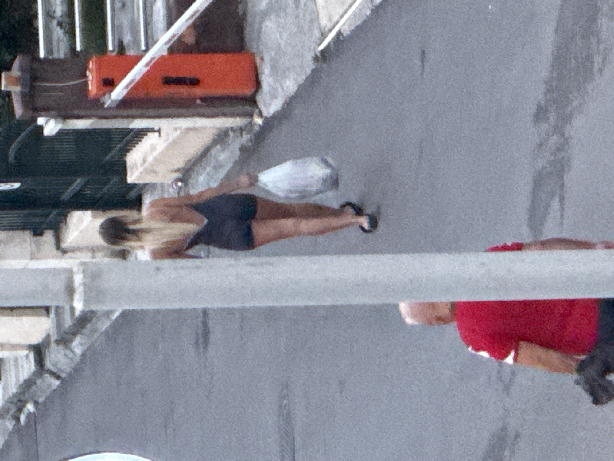 In Budva, Montenegro, a woman with blonde hair pulled back walks away from the camera, wearing a dark, sleeveless, short dress and dark flip-flops. She carries a white plastic bag in her right hand. A man in a red t-shirt with white accents on the sleeves is walking towards the camera in the foreground, his back mostly turned. He appears to be holding a dark object, possibly a leash, and is positioned to the left of a large, light-colored concrete pillar that partially obstructs the view. The scene takes place on a paved street with a slight incline. To the right, a red automatic gate barrier arm is visible, and behind it, a dark metal fence and some vegetation. The overall lighting suggests it is daytime.