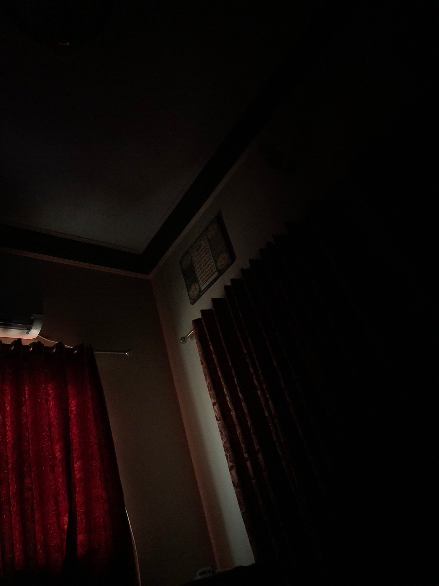 The image shows a dimly lit room, possibly a bedroom, in Larkana, Pakistan. The scene is characterized by the interplay of light and shadow. A red curtain on the left is illuminated, revealing a floral pattern. To the right of it, a white air conditioner unit is partially visible. Further to the right, another red curtain is drawn, mostly obscured by darkness. On the wall, a framed artwork, possibly religious calligraphy, is illuminated by a light source from the right, casting a soft glow on the wall. The ceiling is dark, with a corner molding visible. The overall impression is one of a quiet, private space at night.