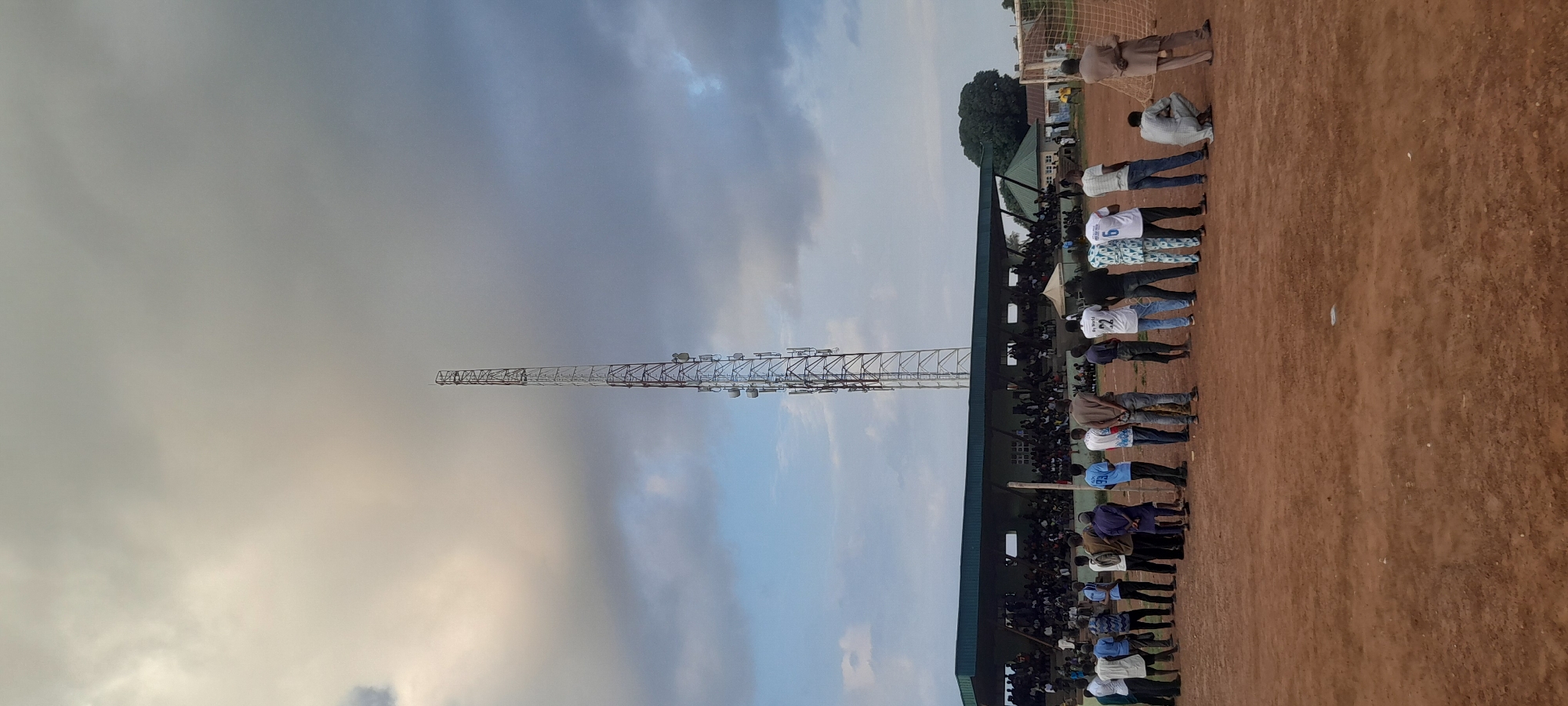 In Abuja, Nigeria, a crowd of people stands in a line on a dirt field. Behind them, a stadium with spectators is visible, along with a tall communication tower reaching into the cloudy sky. The scene suggests a sporting event or gathering taking place outdoors.