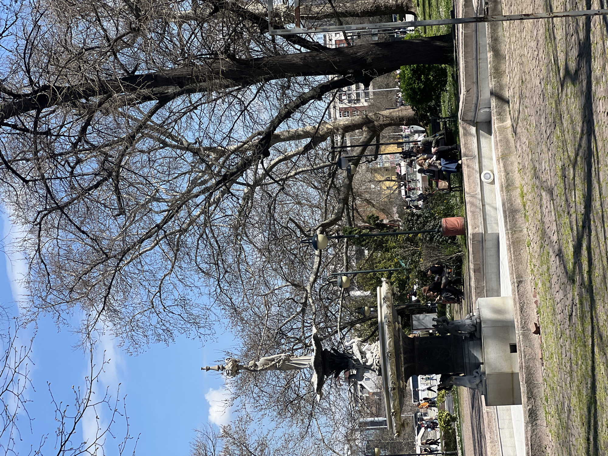 The image shows a public square in Plovdiv, Bulgaria, featuring the Stefan Stambolov monument. The statue, depicting a woman holding a sword, stands atop a multi-tiered fountain. Bare trees cast shadows on the square. People are sitting on benches near the fountain, and others are walking by. The ground is paved with cobblestones. Buildings are visible in the background. The sky is blue with some clouds.