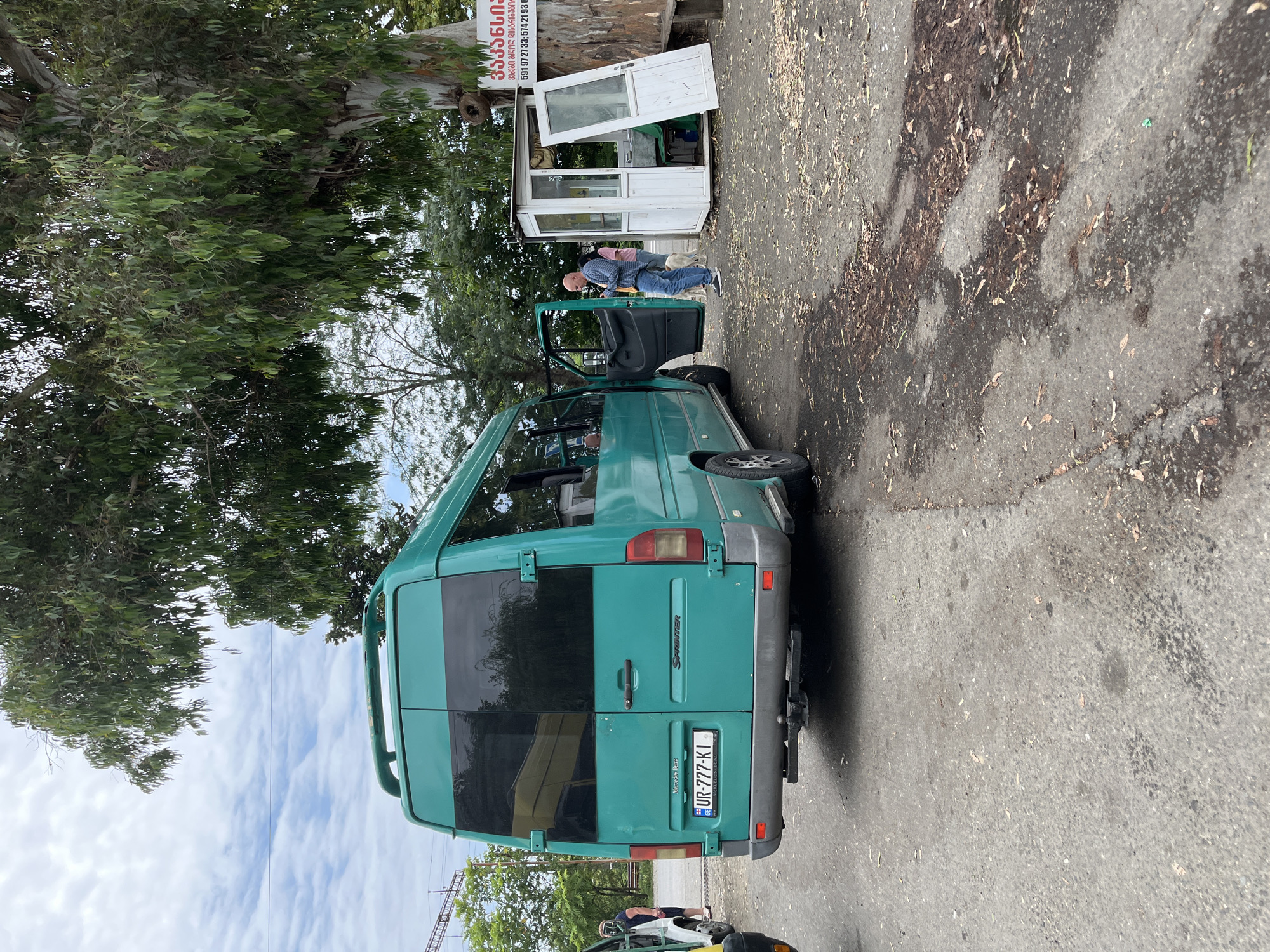 The image depicts a teal-colored Mercedes-Benz Sprinter van parked on a paved surface littered with fallen leaves. The van's rear doors are closed, and its license plate reads "UR-777-KI". To the right of the van, a small white kiosk with an open door stands beside a large tree. A man in blue jeans and a checkered shirt is standing in front of the kiosk, looking down at his phone. A woman in a pink shirt and a white dog are also visible near the kiosk. The background shows a cloudy sky and lush green trees. A sign on the tree next to the kiosk contains Georgian text and a phone number. The scene appears to be a roadside stop in Ureki, Georgia, a resort town known for its magnetic sand beaches.