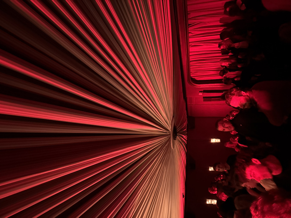 The image depicts an interior scene within a dimly lit venue, characterized by pervasive red ambient lighting. The ceiling is a prominent feature, covered in light-colored fabric drapes arranged radially, converging at a central circular fixture and fanning outwards across the entire overhead expanse. This fabric is heavily saturated with red light, creating a striped effect. On the left wall, several vertical rectangular light fixtures emit a softer, possibly yellowish, glow. To the right, a section of the wall is covered by dense, dark red curtains.
A crowd of people occupies the lower half of the frame, standing and primarily illuminated by the dominant red light. Specific individuals are discernible: a bearded man in the foreground on the right, looking to the left; a man in the mid-ground, slightly left of center, with dark hair, looking directly forward; and a young man with curly hair in the lower left foreground, seen in profile. The individuals appear to be observing or engaged in a social gathering. No specific actions or interactions beyond standing are clearly identifiable.
This scene is located inside Chez Moune, a club situated at 54 Rue Jean-Baptiste Pigalle, Paris, France.