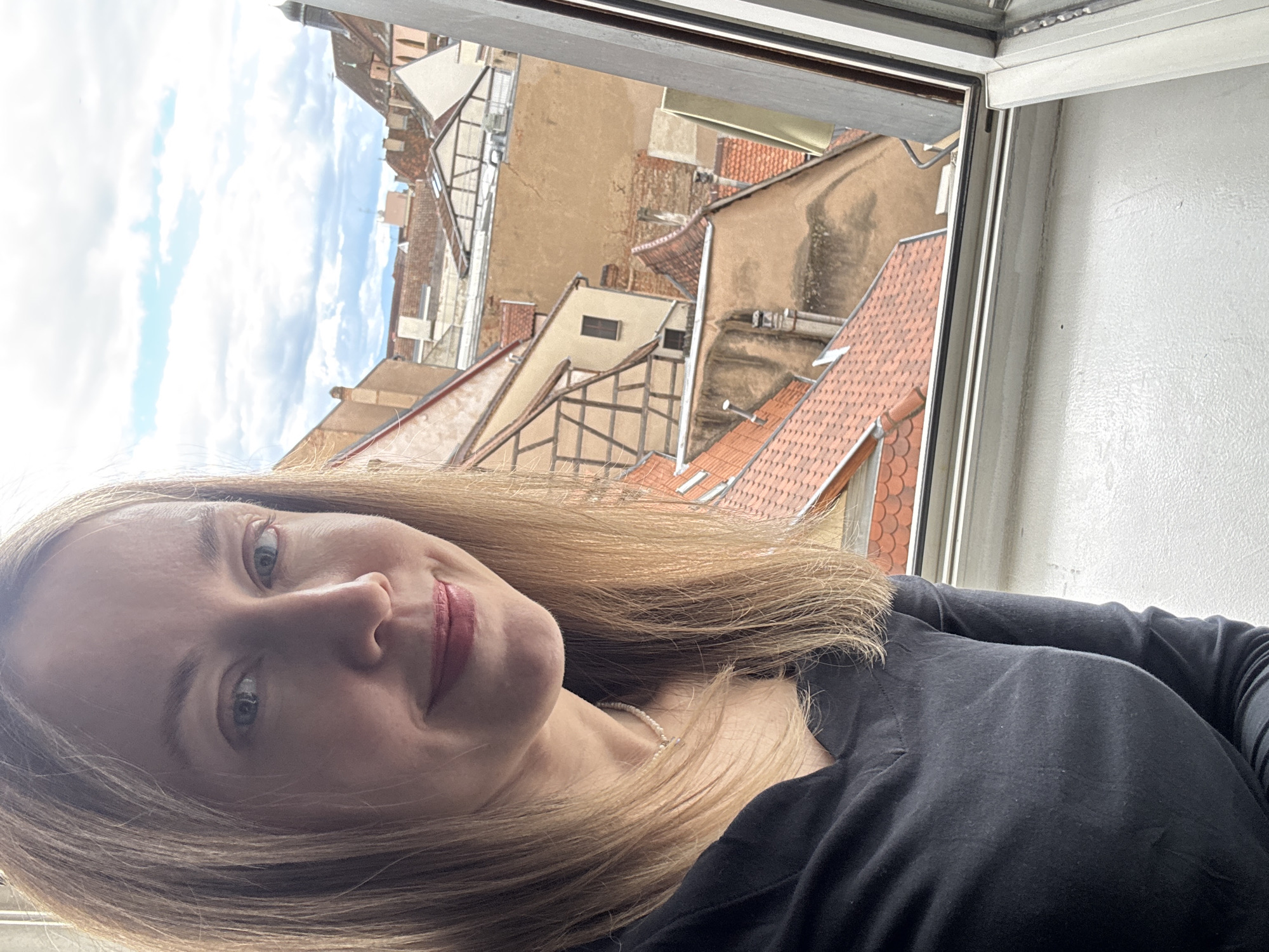 The image shows a medium shot of a Caucasian woman with blonde hair, fair skin, and blue eyes, wearing a dark v-neck shirt and a pearl necklace. She is looking directly at the camera with a slight smile. Behind her, a window reveals a view of the rooftops of Colmar, France. The architecture suggests a historic European city, with terracotta tiles and half-timbered buildings. The sky is partly cloudy. The lighting appears to be natural, coming from the window.