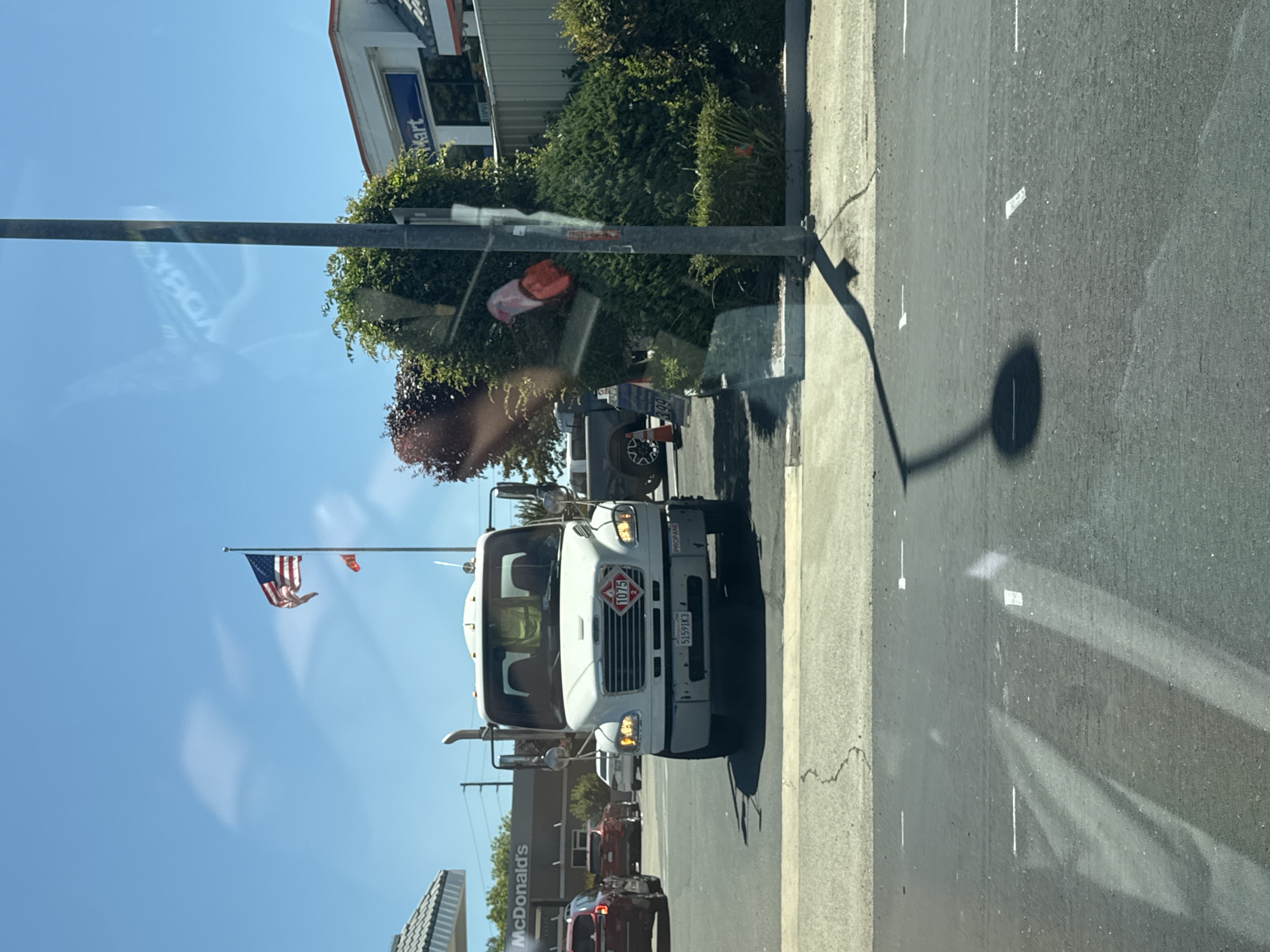 The image captures a frontal view of a white propane truck parked on a street in Citrus Heights, California, on a sunny day. The truck is equipped with a visible exhaust pipe and displays a "1075" hazard symbol on its front grille, indicating it carries flammable gas. A license plate reading "51591K3" and the word "PROPANE" are visible on the truck's bumper. In the background, a McDonald's restaurant and a convenience store are identifiable. A tall pole with a shadow stretches across the pavement. Several vehicles are present in the scene, including a red SUV, another car, and a dark-colored truck. The environment appears to be an urban or suburban setting, with paved roads and sidewalks.