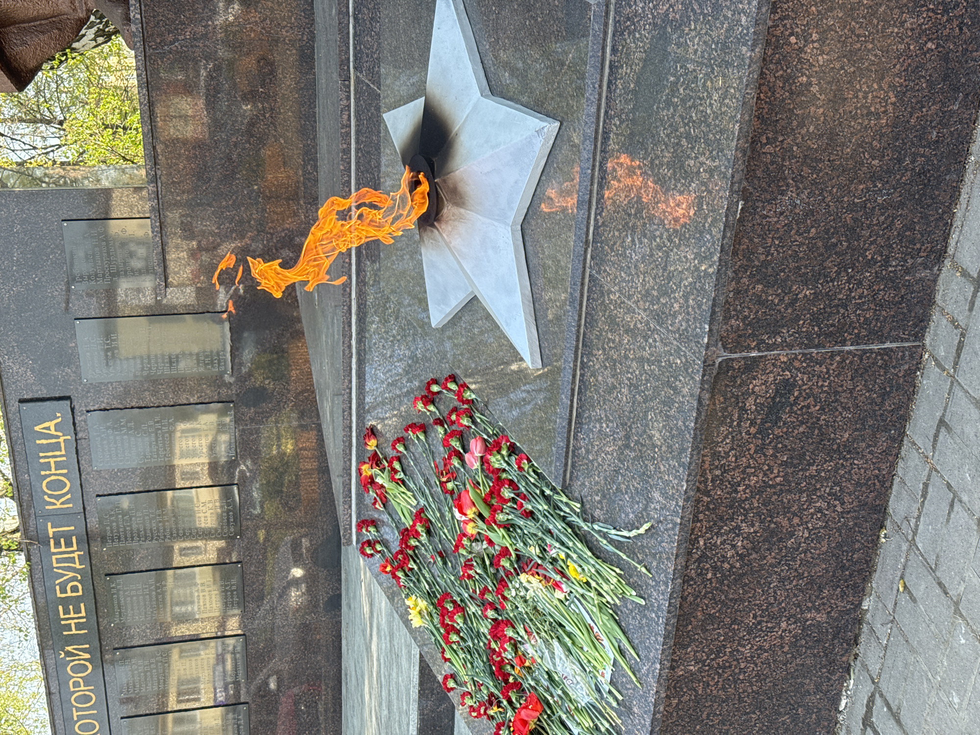 The image shows a war memorial, likely in the city of Zav'yalovo, Russia. It consists of a dark granite wall with bronze plaques containing inscriptions. A five-pointed star with a flame emanating from its center is mounted on the wall. A bouquet of red and yellow flowers rests at the base of the star. The text on the wall reads, "ОТОРОЙ НЕ БУДЕТ КОНЦА," possibly indicating an eternal flame concept.