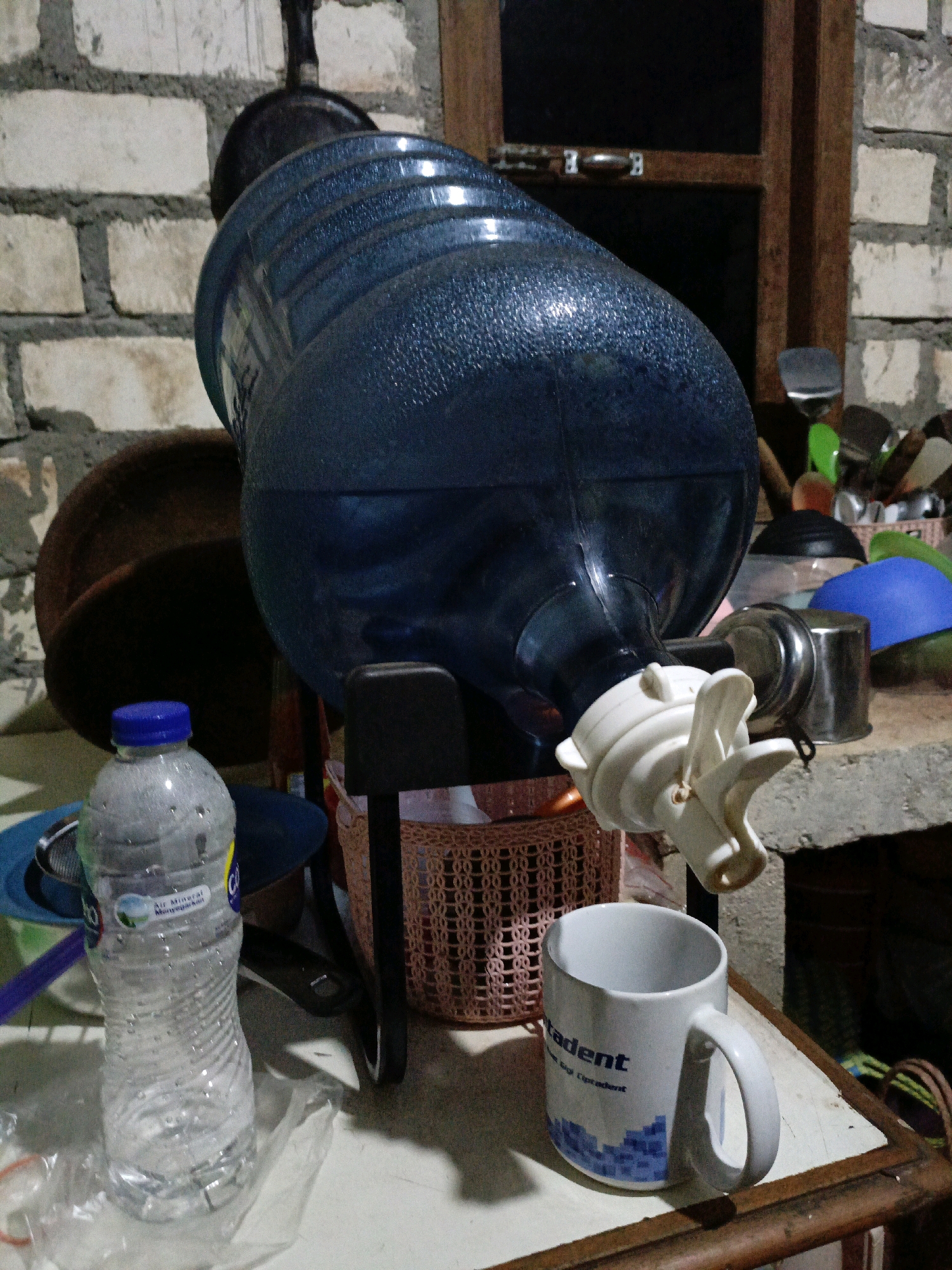 The image depicts a domestic setting, likely a kitchen or storage area, with a large blue water dispenser prominently featured. The dispenser is tilted, with its tap extended downwards. To the left of the dispenser is a partially filled plastic water bottle with a blue cap. In the foreground, a white mug with blue geometric patterns and text that reads "tudent" is placed on a wooden surface. Behind the water dispenser, there are various household items, including a woven basket, a metal canister, and what appears to be a blue lid. The background consists of a brick wall, suggesting the location is Jenu, Indonesia. The lighting in the image is dim, creating shadows and highlighting the textures of the objects.