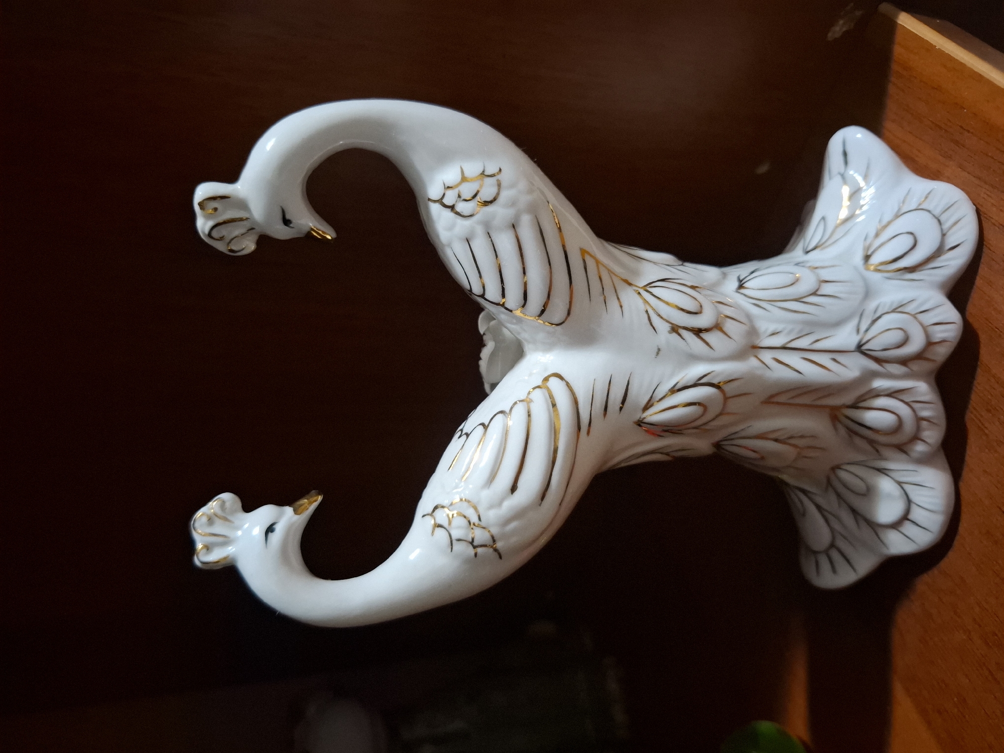 A white ceramic decorative piece shaped like two peacocks facing away from each other. The peacocks have gold accents on their crests, beaks, and feathers. The base of the piece is also decorated with gold feather patterns. The object is placed on a wooden surface, likely a shelf, in a dimly lit room. The background is dark, suggesting a cabinet or a wall. The image quality is moderate, with some graininess. The location is specified as Almaty, Kazakhstan, but no specific landmarks or environmental cues confirm this in the image itself.