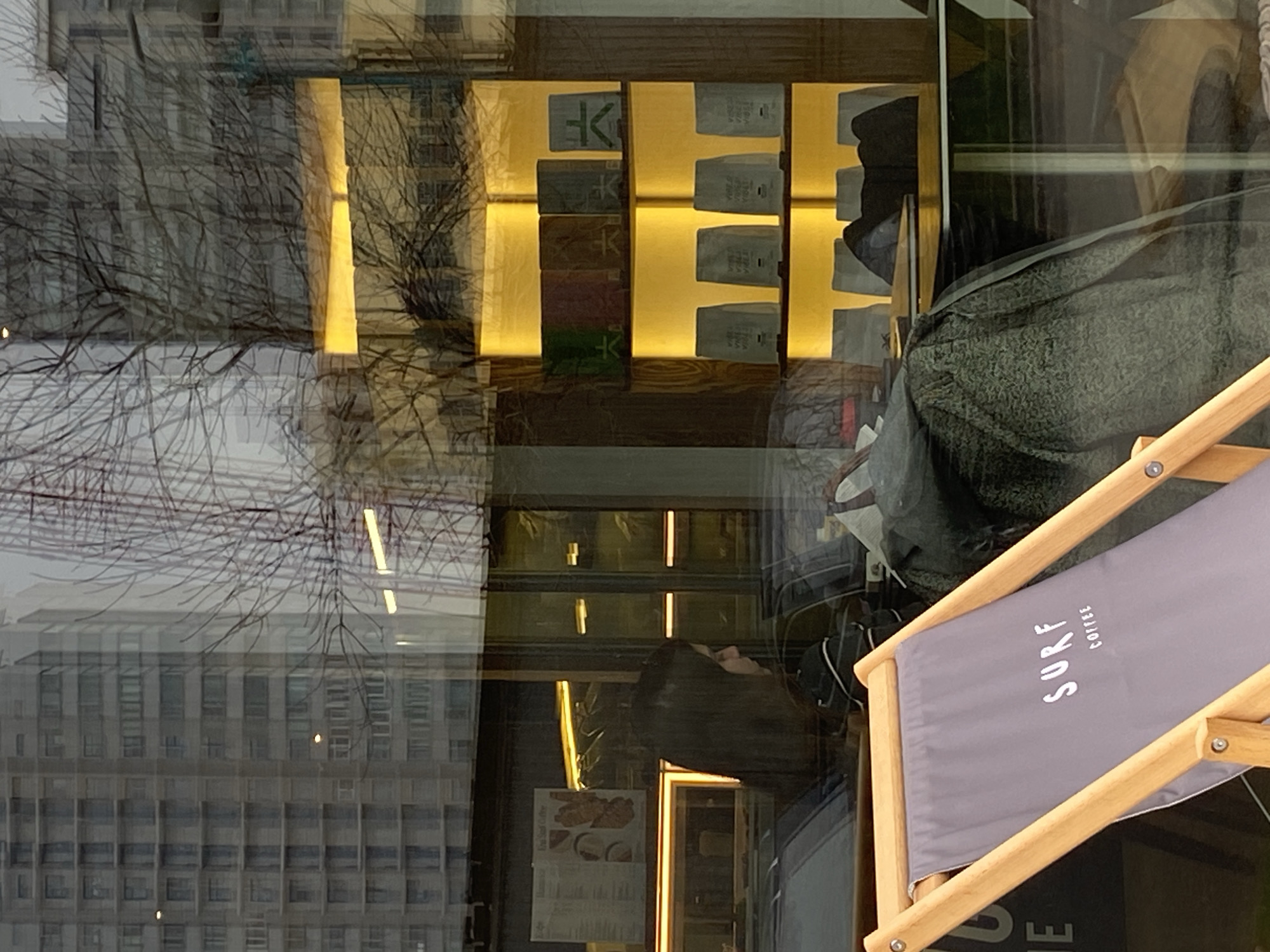 This portrait-oriented image captures a layered scene through the window of a contemporary cafe or shop. In the foreground, positioned in the lower right, is a light wooden-framed folding chair with a grey fabric seat. The fabric bears the white text "SURF COFFEE".
The glass window acts as a mirror, reflecting a complex urban exterior. Bare tree branches, intricately silhouetted against an overcast, grey sky, suggest a colder season like winter. Behind the branches, the reflection of a large, multi-story building with numerous grid-patterned windows is prominently visible.
Inside the establishment, a warm ambiance is created by horizontally arranged shelves with bright yellow backlighting. These shelves display various product packages or boxes. Some white packages clearly show the word "VIBES," while other colored packages (green, brown, grey) feature a distinct, stylized character, possibly an East Asian symbol or a brand logo.
Faintly visible further inside the shop is the reflection of a person with long dark hair, positioned near a wall where a menu or poster is displayed. A dark, textured fabric, possibly a blanket or part of an interior furnishing, and a large dark bag are also discernible, partially obscured by the reflections. The scene depicts a modern retail space within an urban setting, consistent with a city environment like Moscow, Russia.