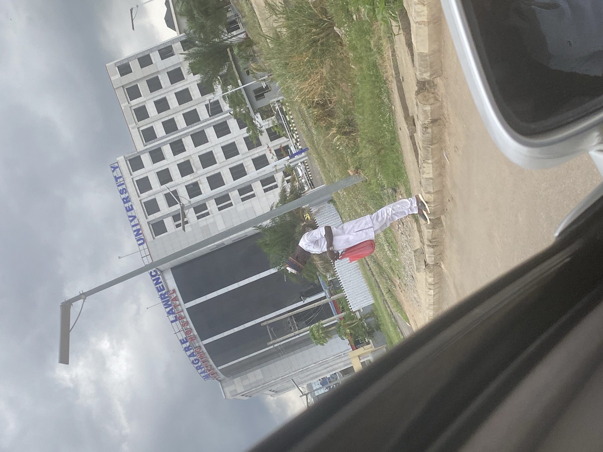 The image shows a view from inside a car, likely parked on the side of a road in Abuja, Nigeria. On the left side of the image is the Margaret Lawrence University Teaching Hospital. A man wearing a white outfit and a red bag or parcel is standing on the sidewalk. The sky is overcast, and the environment appears to be urban with some greenery. The view is partially obstructed by the car's interior.