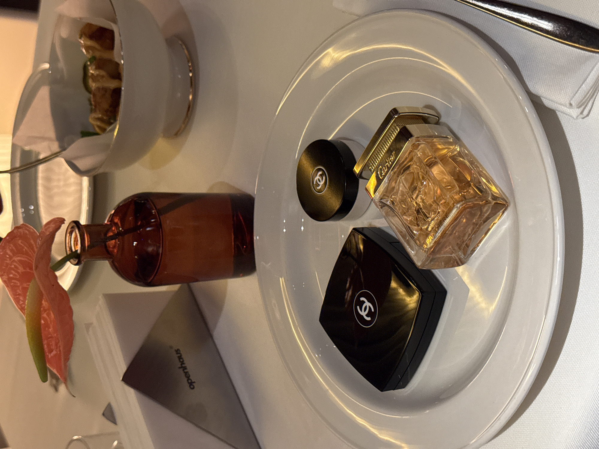 The image displays a close-up, elevated view of a dining table at Openhaus, a establishment located in Yerevan, Armenia. The table is covered with a white tablecloth. In the foreground, a white circular plate holds three items: a rectangular black compact featuring a white Chanel logo, a small circular black-lidded container with a white Chanel logo, and a clear glass perfume bottle with gold-toned accents, labeled "Cartier," containing an amber liquid. To the right of the plate, a silver fork and a white folded napkin are partially visible. In the midground, an amber glass vase contains a single pink anthurium flower with a green and yellow stem. Adjacent to the vase, a white, triangular napkin holder displays the "openhaus" logo in black text. In the background, a white bowl with a gold rim holds several food items, appearing to be wrapped or served in white paper, and topped with a light-colored sauce. A blurred clear glass and another white plate are also visible. The scene is illuminated by warm, artificial light. No individuals are present.