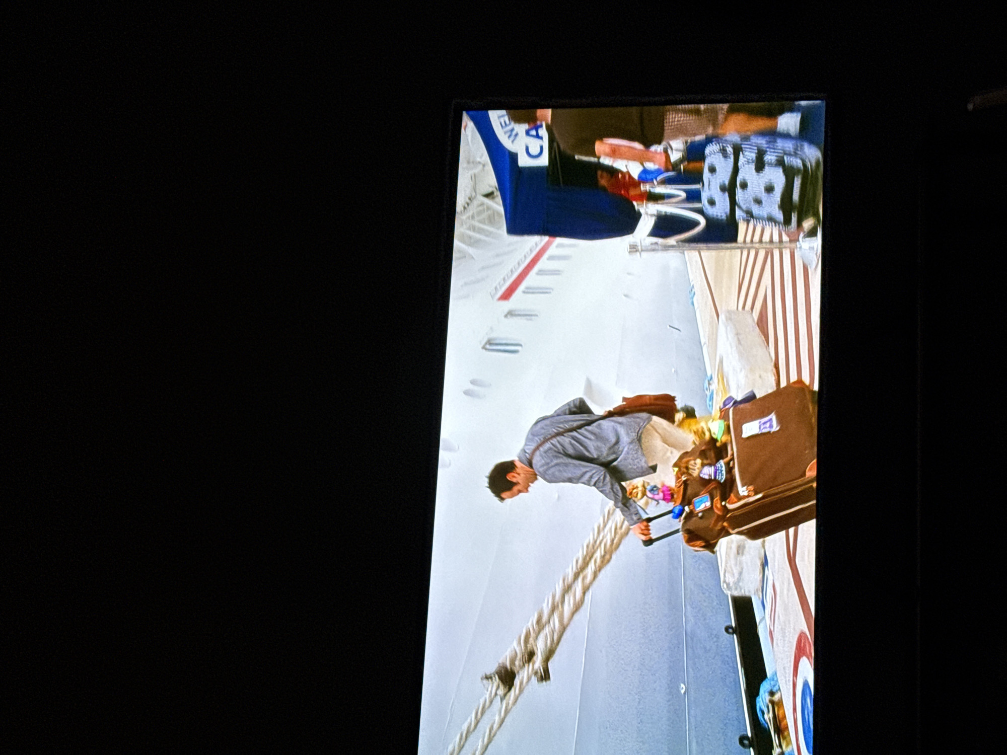 The image shows a television screen displaying a scene depicting a man disembarking from a cruise ship. The man is dressed in a gray jacket and light-colored pants. He is pulling two brown suitcases up a gangplank from the ship. The cruise ship has the word "Carnival" visible on the side. The surrounding environment appears to be a sunny day. There is no indication of this being in the city of College Station, United States of America.