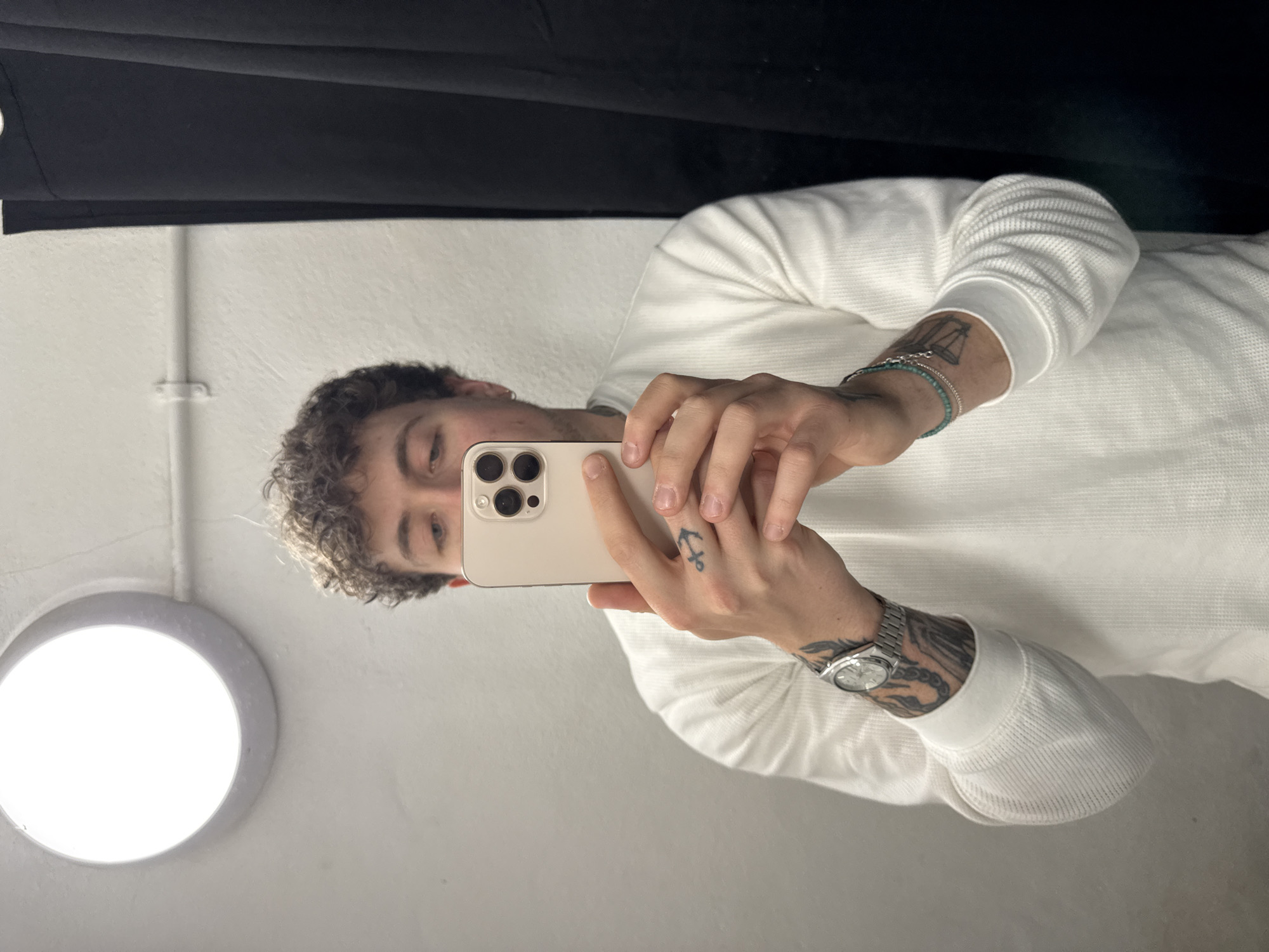 A young man is depicted taking a mirror selfie indoors. He is seen from the chest up, with his head tilted slightly and eyes looking down towards the light-colored smartphone held in his hands. He has curly dark hair and is wearing a white, long-sleeved, waffle-knit shirt.
On his left wrist, he wears a silver chain bracelet and a turquoise beaded bracelet. A small anchor tattoo is visible on the back of his left hand, and a tattoo of the scales of justice is partially visible on his left forearm. On his right wrist, he sports a silver-toned analog watch with a metal band, and an intricate black and grey tattoo covers a portion of his right forearm.
The background features a white wall with a subtle texture. To the left, a bright, circular, recessed light fixture emits white light, illuminating the scene. A thin white pipe or conduit runs vertically along the wall. At the top of the frame, a dark black fabric or material, possibly a curtain or backdrop, is visible. The setting appears to be an interior space, such as a fitting room or a room with minimalist decor. The lighting is artificial and bright. There are no specific cues for the time of day or weather, and the man's expression is neutral. No text is visible in the image. The image is from London, United Kingdom.