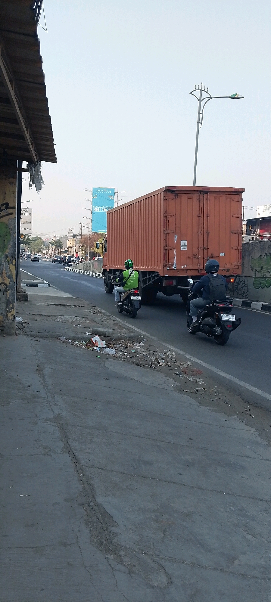 The image depicts a street scene in Bekasi, Indonesia. In the foreground, a damaged concrete sidewalk is covered in debris. The sidewalk abuts a road where traffic is moving. A large orange truck with a closed trailer is the dominant vehicle in the scene, traveling away from the viewer. Two individuals on motorcycles are also visible, following the truck. One motorcyclist, wearing a green jacket with a Gojek logo, is positioned to the left of the truck. The other motorcyclist, wearing a blue backpack and a dark helmet, is to the right of the truck. The road is bordered by a curb and a white line. In the background, there are buildings, streetlights, and a large blue billboard. The sky is clear and light blue.