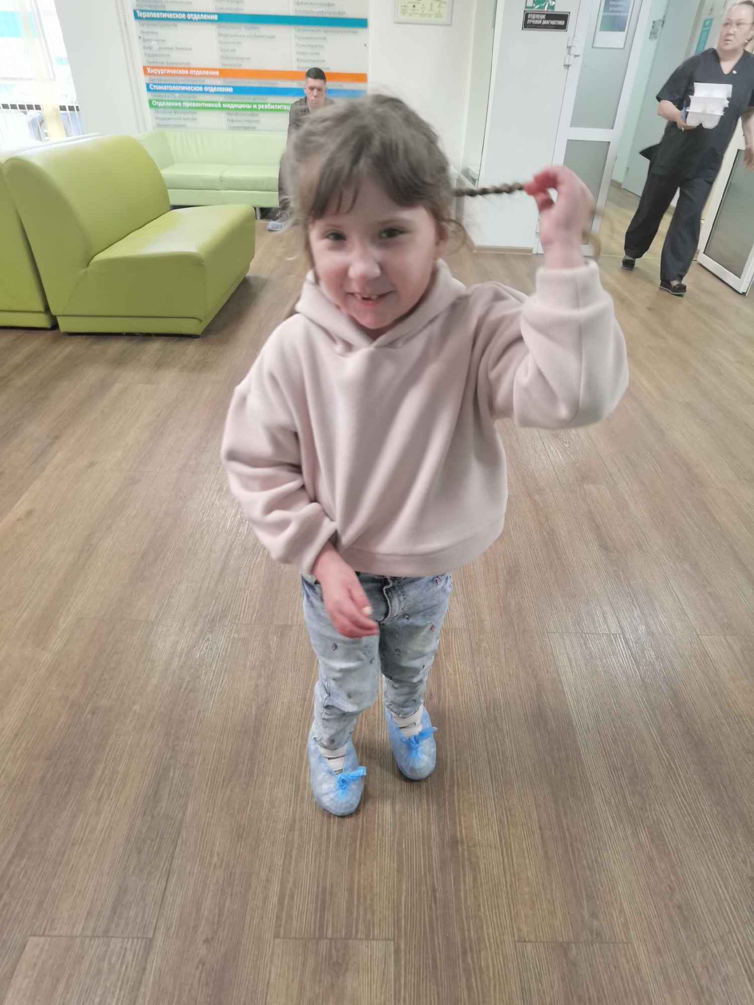 The image captures an indoor scene within a medical facility, located in Izhevsk, Russia.
In the foreground, a young girl, estimated to be 3-5 years old, stands centrally, facing the camera with a broad smile. She has light brown, curly hair styled into two small braids, one of which she holds with her right hand, lifting it slightly. She is dressed in a light pink, long-sleeved hooded sweatshirt and light blue jeans adorned with small, colorful embroidered or printed details. Her feet are covered in light blue disposable shoe covers, secured with blue ribbon ties.
The flooring consists of light brown wood-effect laminate or vinyl planks. The walls are white. To the left of the frame, two bright lime-green upholstered modular benches are arranged, forming part of a waiting area. Seated on one of these benches in the mid-ground, an adult male wearing a dark top and dark pants is visible, partially obscured by the girl's head and facing away from the camera.
On the back wall, a prominent blue-and-white sign in Russian lists various departments: "Терапевтическое отделение" (Therapeutic Department), "Хирургическое отделение" (Surgical Department), "Стоматологическое отделение" (Dental Department), and "Отделение превентивной медицины и реабилитации" (Department of Preventive Medicine and Rehabilitation).
In the background, on the right side of the image, an adult female wearing dark medical scrubs and glasses is in motion, walking towards the right. She holds a white tray containing multiple small white cups or containers in her right hand. Above a white door behind her, a smaller white sign with a green arrow indicates "ОТДЕЛЕНИЕ ЛУЧЕВОЙ ДИАГНОСТИКИ" (Department of Radiation Diagnostics).