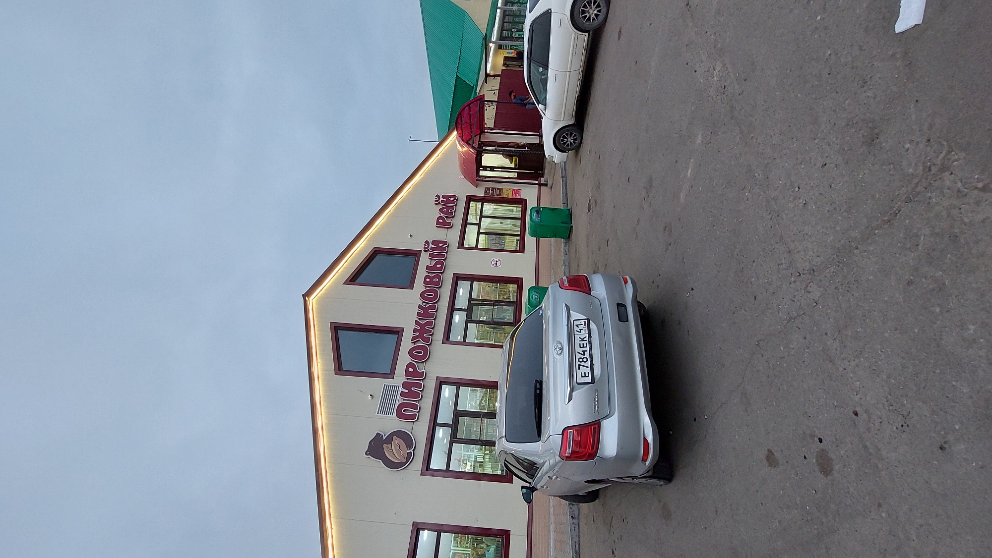 The image depicts the exterior of a commercial building in the city of Sokoch, Russia. The building, which appears to be a store, has a beige facade with prominent red lettering that reads "ПИРОЖКОВЫЙ РАЙ" (Pirozhkovy Ray), translating to "Pie Paradise." Above the lettering, a logo featuring a bear holding what looks like a pastry is visible. The building has several large windows and a pitched roof, with decorative lighting along the eaves.
In the foreground, a silver Toyota sedan is parked on an asphalt lot. The license plate is visible and reads "E784EK 41". To the right of the Toyota, a white sedan is parked. Two green trash receptacles are positioned near the building's entrance. The sky is overcast, suggesting a cloudy day. The overall environment is that of a typical commercial area in a Russian city.