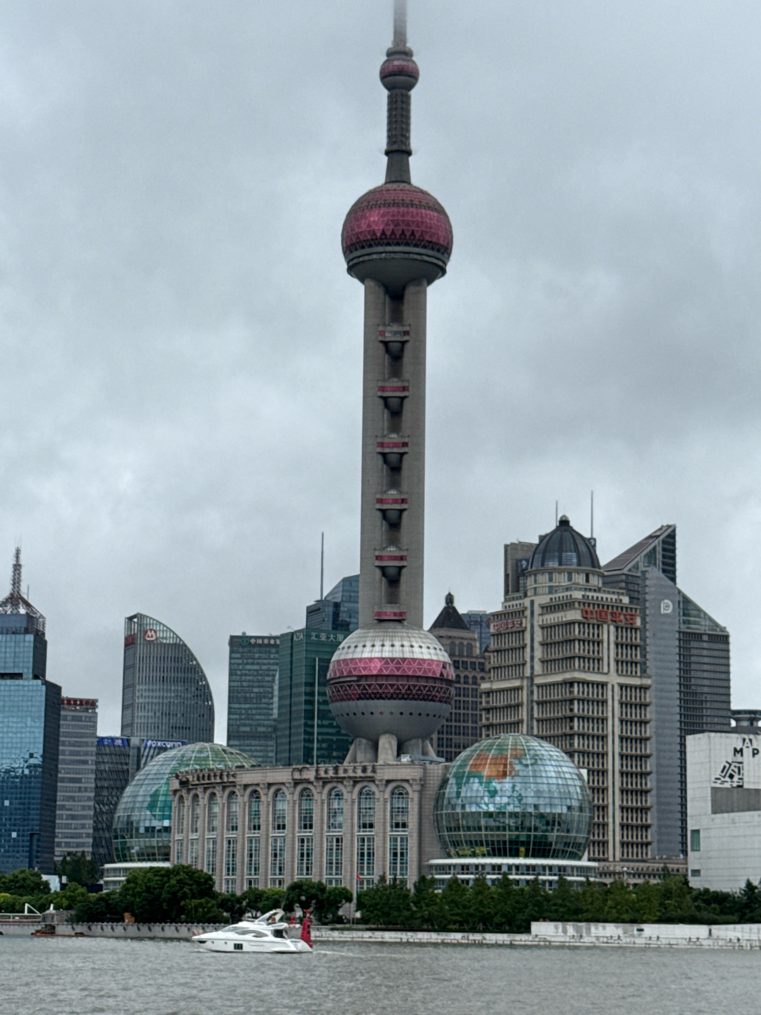 The image depicts the Shanghai skyline, prominently featuring the Oriental Pearl Tower. The tower is a tall, multi-tiered structure with spherical elements, two of which are accented with pink and red designs. Surrounding the tower are various modern skyscrapers, characterized by their glass facades and diverse architectural styles. In the foreground, a body of water, likely the Huangpu River, is visible, with a white yacht cruising on its surface. Green trees line the riverbank, interspersed with buildings and pathways. The sky is overcast, contributing to a muted color palette. The overall scene captures a view of Shanghai's Pudong district, known for its distinctive modern architecture and iconic landmarks.