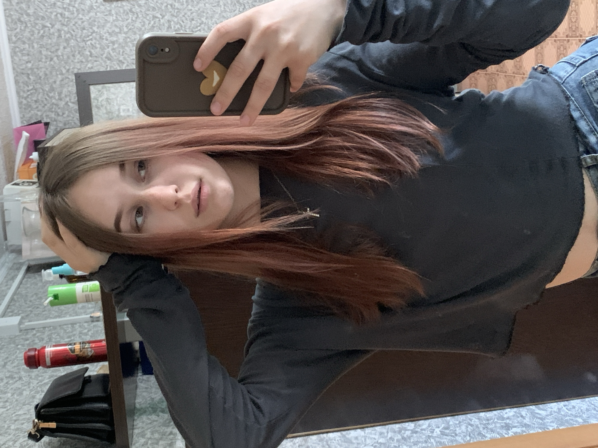 A woman is taking a selfie with a brown phone with a heart-shaped sticker on the back. She is wearing a black long-sleeved shirt and jeans. She has long, brown and blonde hair. A black purse, a red bottle, and green and blue spray bottles are visible on a shelf behind her. The location is not discernible as being in the city of Фоки, Russia.