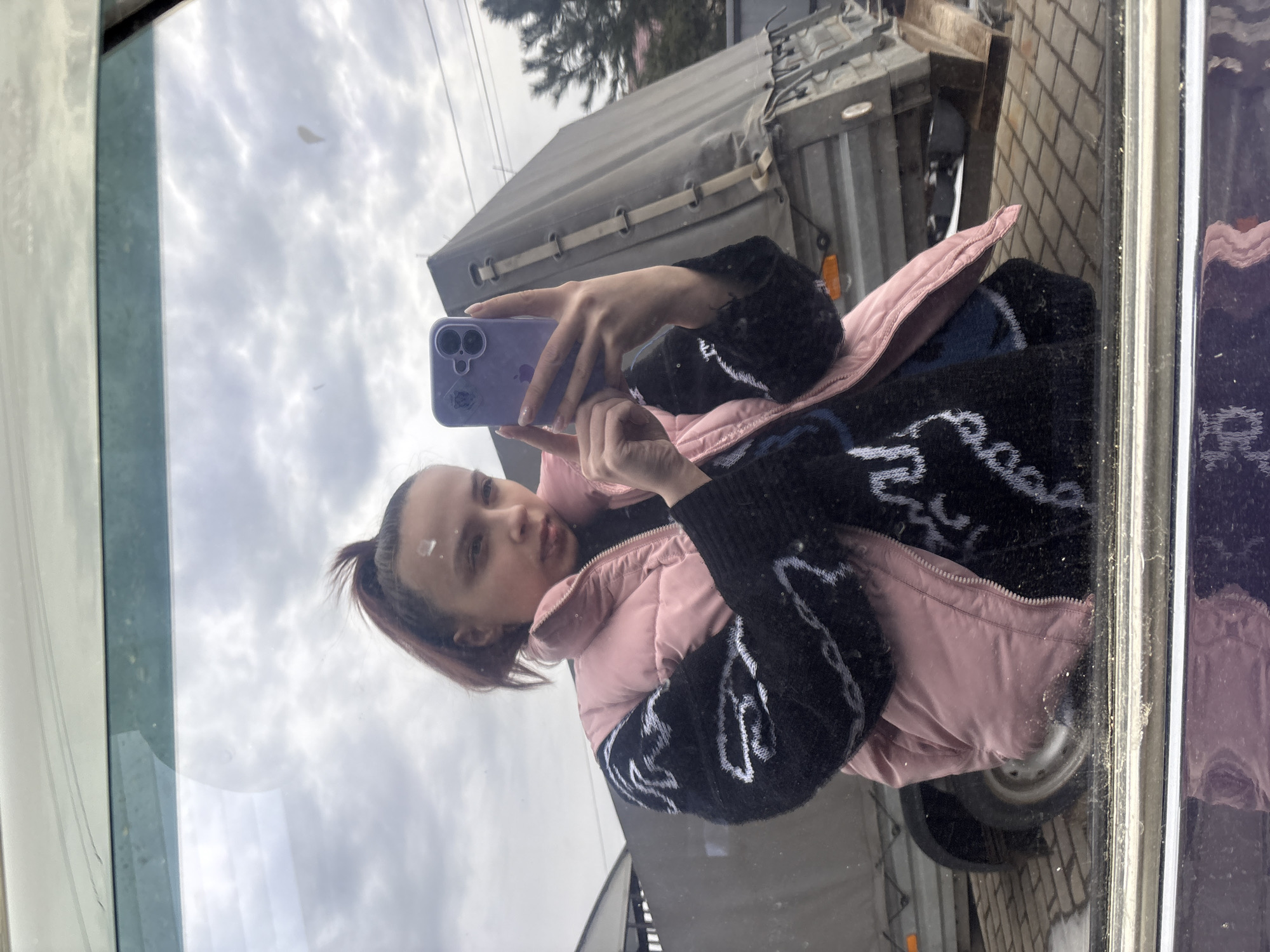 A young woman is taking a selfie with a purple smartphone in a reflective surface. She wears a pink vest over a black sweater with white designs. The reflection shows a trailer. The location is Прикамский, Russia.
