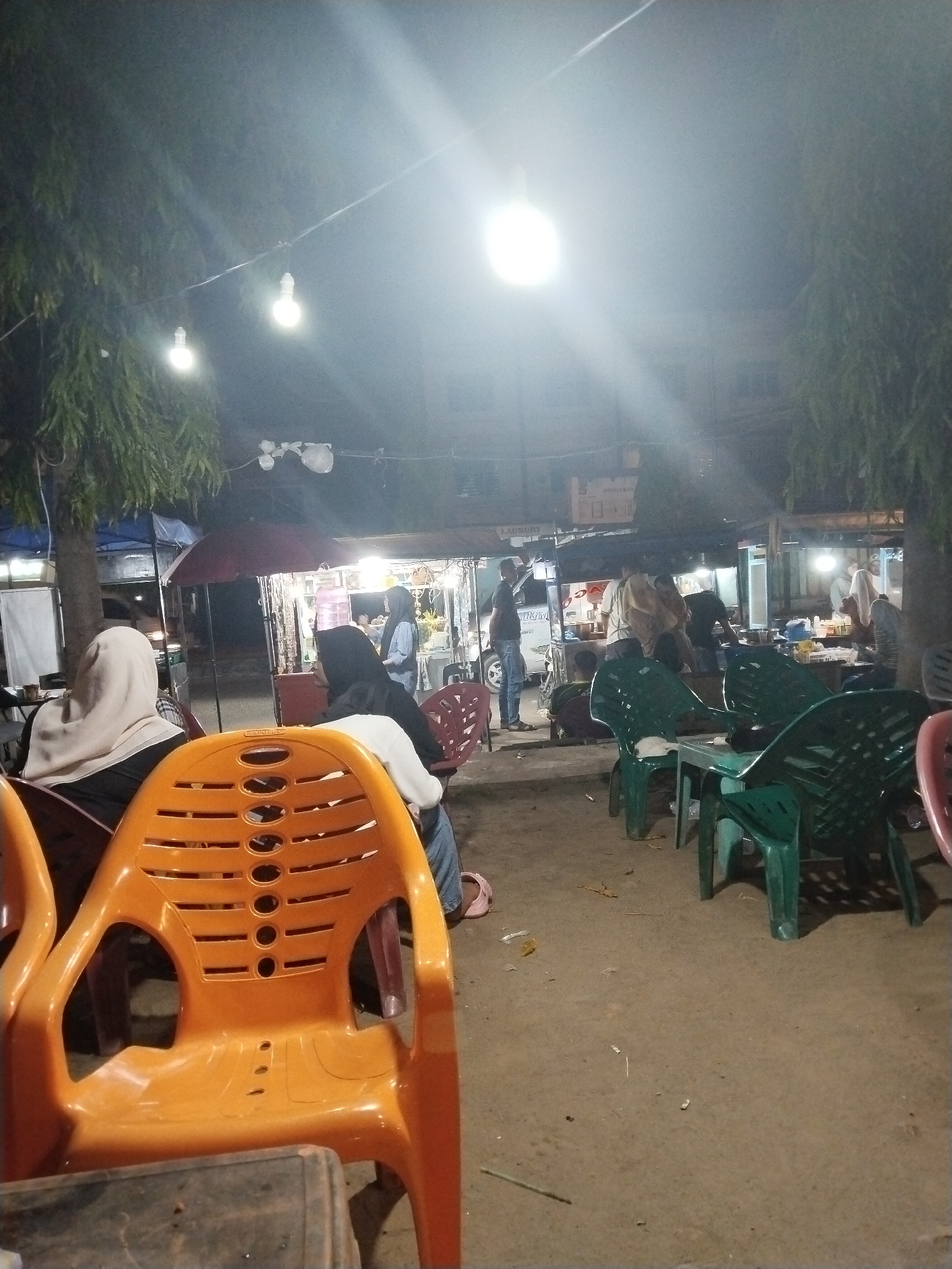This is a nighttime photograph of an outdoor eatery in Lhokseumawe, Indonesia, likely a street-side food stall or warung. Several people are present, some seated at tables, and others appear to be employees or customers at the food stalls in the background. The scene is illuminated by hanging light bulbs, casting a bright, somewhat hazy light.
In the foreground, an orange plastic chair is prominently displayed, with another chair partially visible to its left. The ground is a dirt or gravel surface.
In the middle ground, several people are seated on plastic chairs, some wearing headscarves. In the background, there are food stalls with awnings and lighting. One stall has a sign that reads "LAUNDRY". People are seen preparing food or serving customers. There is a white vehicle parked to the left.
The overall atmosphere is informal and typical of a casual dining spot in Southeast Asia. The presence of trees on either side suggests the location might be along a street or in a small open area within the city.