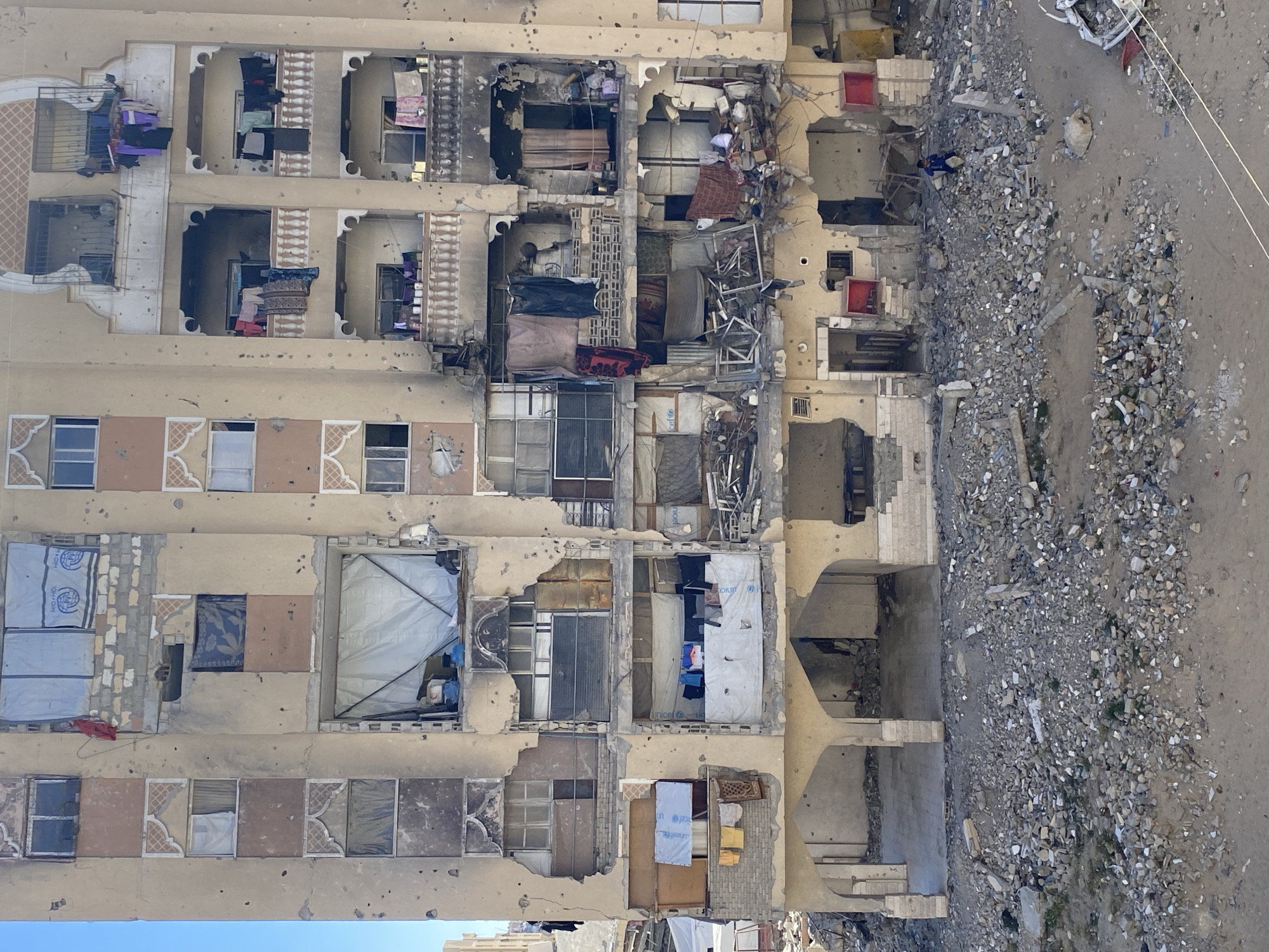 The image, rotated 90 degrees counter-clockwise, depicts a severely damaged multi-story residential building, likely an apartment complex, bordering a wide, rubble-strewn street or alleyway. The structure shows extensive signs of destruction, including large sections of walls blown out, exposed interiors, shattered window frames, and numerous pockmarks on its facade consistent with bullet holes or shrapnel damage.
Inside the exposed rooms, remnants of daily life are visible amidst the devastation. Clothes are seen hanging to dry on makeshift lines on balconies and within damaged window openings, suggesting a continued or recent human presence. Bedding, furniture fragments, and general household debris are strewn within the gutted rooms. White and blue tarpaulins, some bearing the UNICEF logo, are used to cover openings, serving as makeshift walls or temporary shelters within the damaged structure.
The ground adjacent to the building is covered in a dense layer of concrete chunks, rebar, bricks, and other construction debris, indicating a significant collapse or impact. In one area of the rubble, a small figure dressed in blue clothing is visible, possibly a child or adult surveying the damage. Several red, rectangular objects, possibly large containers, are scattered on the ground and within the lower levels of the destroyed sections. Overhead, what appear to be power lines sag low across the scene. The sky, visible at the bottom of the main image, is clear blue, indicating daytime. The scene conveys a strong sense of devastation and the aftermath of conflict or a major disaster.