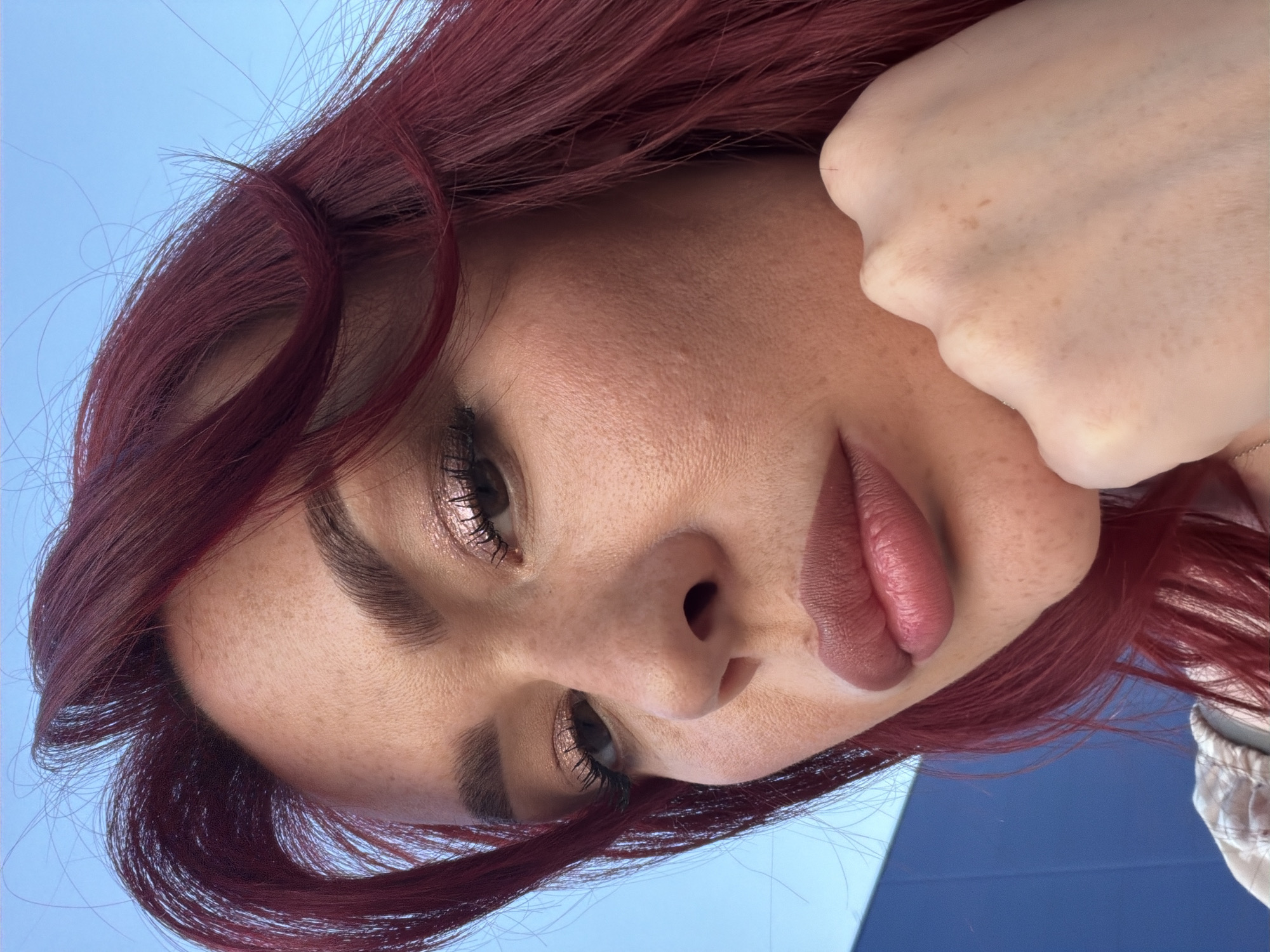 The image is a close-up, low-angle shot of a woman with red hair and fair skin with freckles. She is wearing makeup, including eyeshadow and lipstick. Her right hand is resting on her chin, and her eyes are looking towards the camera. The background is a bright blue sky and a section of a blue building. The scene appears to be outdoors, possibly in a city, but no specific landmarks are visible to confirm a location in Tolyatti, Russia.