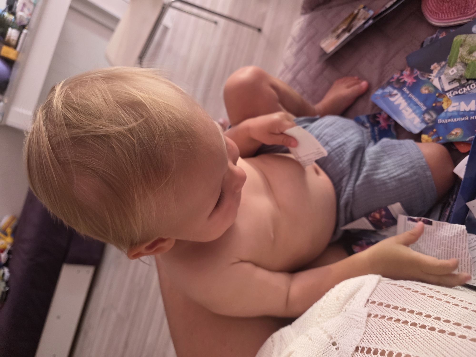 This is a close-up, high-angle shot of a young child, likely a toddler, with blonde hair and fair skin, lying down on their side. The child is wearing light blue shorts and is shirtless, showing their stomach and chest. They are holding a small, white piece of paper in their right hand.
The child is situated on a surface that appears to be a bed or couch, covered with a textured, dark fabric. In the background, there are several items that look like books or game cards, some with colorful illustrations. One card clearly shows the word "MEMO" in large letters, and another has text in Russian that includes "КОСМО" (likely part of a word related to space) and "Подводный мир" (underwater world). The overall setting suggests an indoor environment, possibly a living room or bedroom.
The lighting is soft and diffused, indicating it could be daytime, but the exact time of day is not discernible. There are no visible cues related to weather. The child's expression is not clearly visible due to the angle and focus. The presence of children's books or games suggests a domestic, family setting.
The background also shows a hint of furniture, possibly a closet or a shelf unit, and a purple-covered object, potentially a cushion or part of a sofa. The floor appears to be made of light-colored wood or laminate. The child's left arm is tucked in, and their legs are bent at the knees, with their feet visible in the upper right portion of the frame. The overall mood conveyed is one of quiet play or rest.