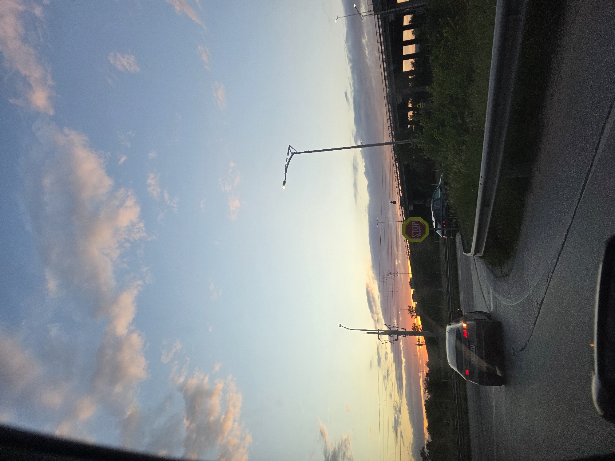 The image depicts a road scene at dusk, likely taken from inside a moving vehicle. The sky is visible, exhibiting hues of blue, pink, and orange, suggesting either sunrise or sunset. A car with its rear lights illuminated is positioned on the right side of the road. There is a yellow stop sign near another parked car. Power lines and streetlights are running parallel to the road. In the background, there is a bridge, potentially a railway bridge, spanning over the road. Given the presence of infrastructure and road traffic, it is probably in Plovdiv, Bulgaria. There are green trees and grass near the bridge.