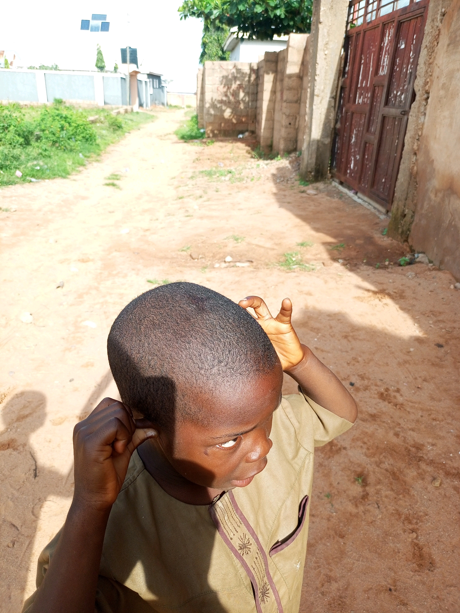 The image depicts a young boy with dark skin and a shaved head, wearing a light brown tunic with embroidered details, standing outdoors on a dirt path in what appears to be a village or suburban area in Tempure, Nigeria. He is looking to his right, with his right hand touching his ear and his left hand raised towards his head. The background includes a dirt road leading away from the viewer, flanked by vegetation on the left and a wall with a brown metal gate on the right. In the distance, there are buildings, including one with solar panels on its roof, and a water tank. The lighting suggests it is daytime, with shadows cast on the ground.
