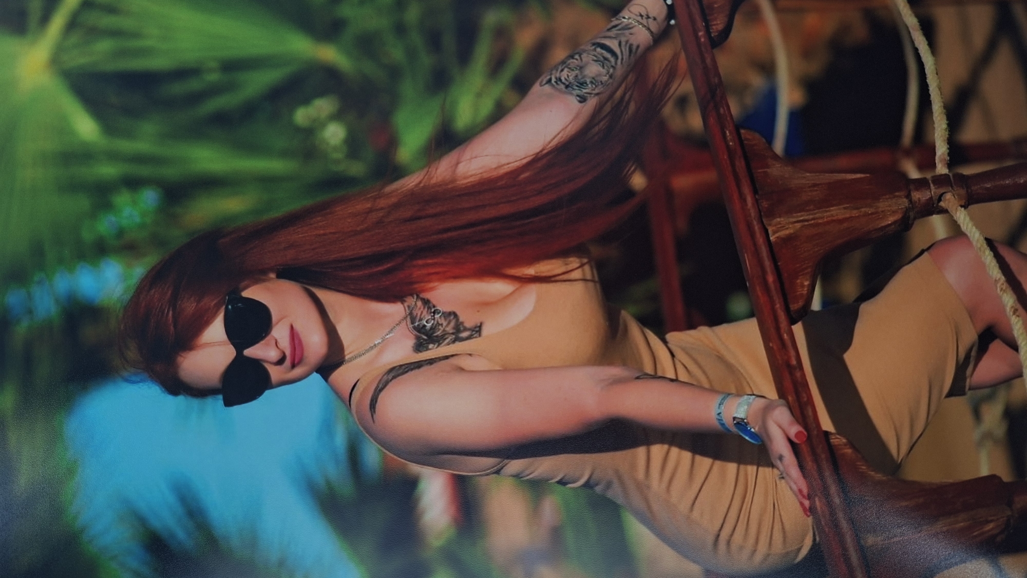A woman with long red hair, wearing dark sunglasses and a tan dress, is photographed in a studio setting. She has multiple tattoos on her arms and chest. The background appears to be a tropical scene, possibly projected onto a screen or wall. She is positioned on a wooden structure resembling a ladder or scaffolding, partially supported by ropes. The location is Забалоцкі сельскі Савет, Belarus.
