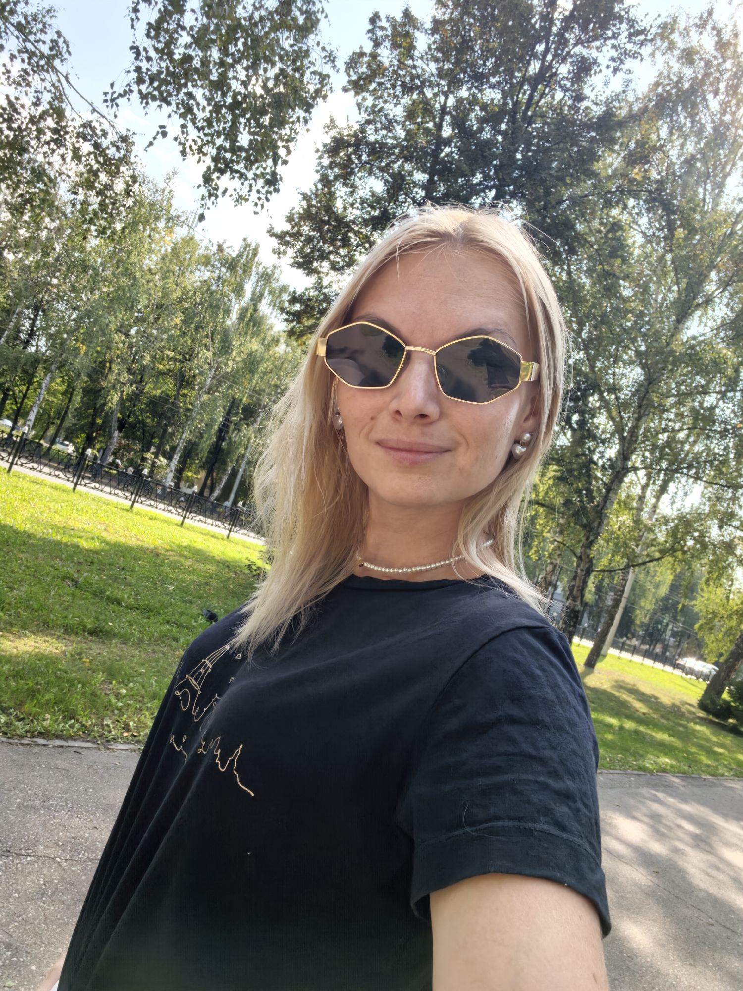 A blonde woman wearing sunglasses and a black t-shirt with gold foil lettering stands in a park on a sunny day. She has a pearl necklace and pearl earrings. Behind her, there are trees and a path. The scene appears to be in the city of Sterlitamak, Russia.