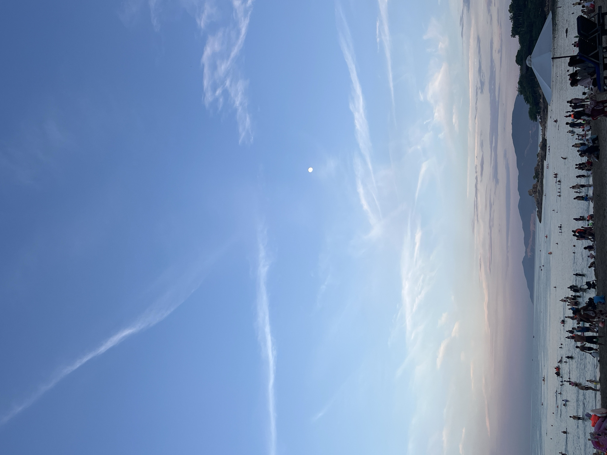 The image depicts a crowded beach scene at twilight in Nha Trang, Vietnam. The sky is a gradient of blue to pale pink, with wispy clouds and a full moon visible. The ocean is filled with people swimming and playing, with small boats scattered across the water. The beach is packed with sunbathers and families. In the foreground, a white umbrella shades two lounge chairs. The background features a rocky island and lush green hills.