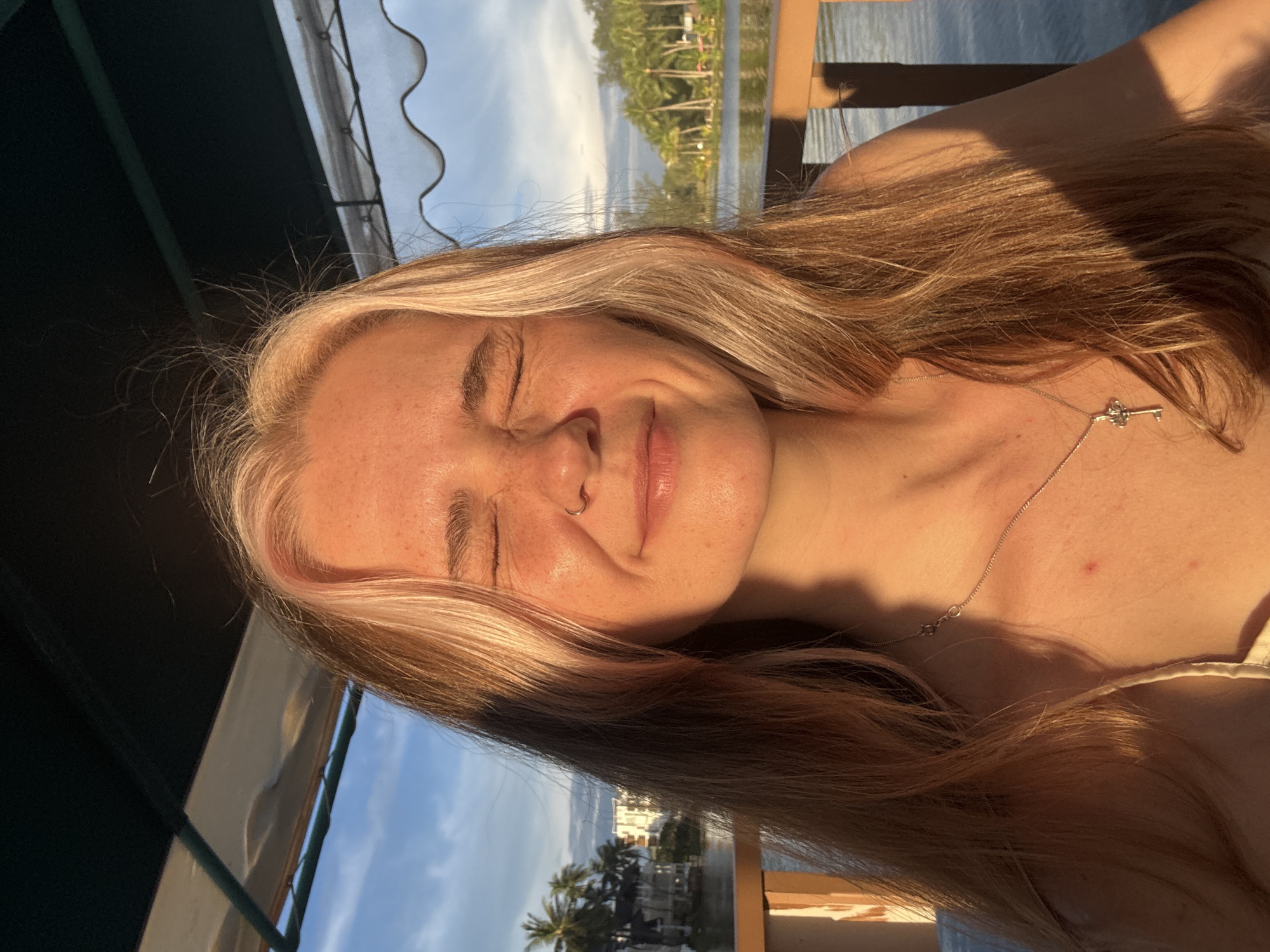 A young woman with blonde and brown hair, wearing a necklace with a key pendant, is pictured with her eyes closed and a slight smile on her face. She is outdoors, likely on a balcony or deck, with a body of water and palm trees visible in the background. The setting appears tropical, consistent with the location of Choeng Thale, Thailand. The lighting suggests it is either sunrise or sunset, casting a warm glow on her skin.