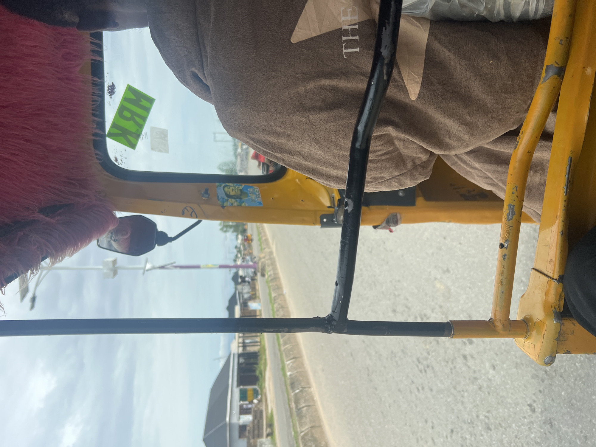 The image captures an interior view from the passenger seat of a yellow auto-rickshaw, possibly a "Keke Napep" or similar, in Jalingo, Nigeria. The perspective is from inside the vehicle, looking outwards and slightly upwards.
The most prominent subjects are the vehicle's interior elements: a pink fluffy material on the left, likely a seat cover or decoration, a part of a person's head and shoulder wearing a brown garment with a light-colored geometric pattern and the word "THE" visible on it, and various metal bars and yellow painted structures that form the auto-rickshaw's frame. A black metal bar, part of the passenger safety structure, runs horizontally across the middle of the image, obscuring the view of the road ahead to some extent.
On the window, a bright green sticker with black lettering is visible. The lettering appears to be "ABK" rotated. Below it, a small sticker with a person's face is affixed to the yellow body of the vehicle.
The scene outside the auto-rickshaw is a somewhat blurred view of a road, with buildings and vegetation lining the side. The sky is visible, appearing overcast or hazy, suggesting it might be daytime with diffused light. The presence of roads, buildings, and the type of vehicle strongly indicate an urban or semi-urban setting in Nigeria.
The image appears to be taken while the auto-rickshaw is in motion, given the slight blur of the exterior scene and the perspective from within a moving vehicle. There are no overt emotional cues, but the general composition suggests a common mode of transport in the region. The overall impression is one of everyday travel in Jalingo, Nigeria.