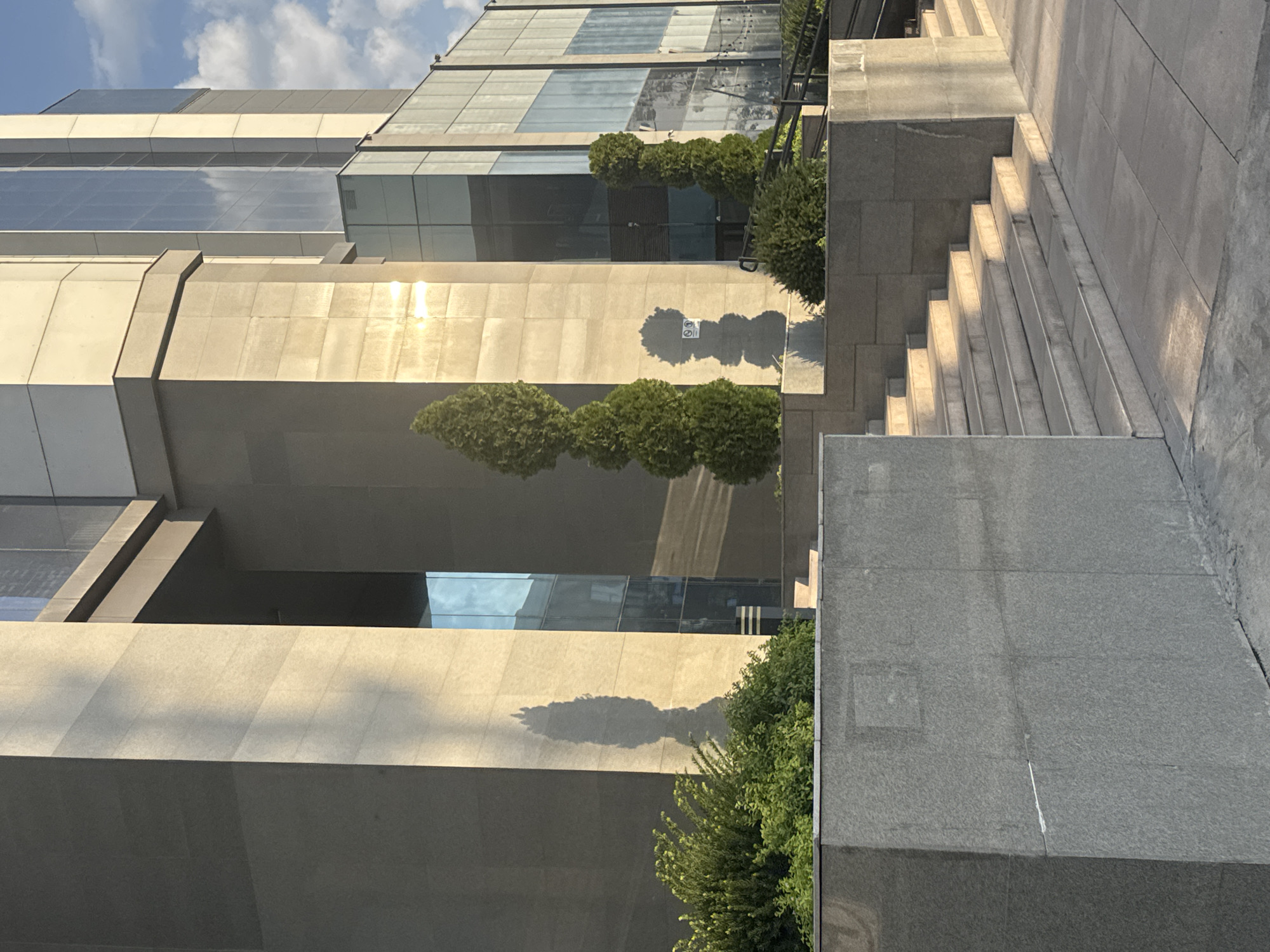 The image captures an exterior view of a modern building with a facade primarily composed of light-colored stone and glass panels. The sunlight casts long shadows across the stone surfaces, indicating it is either early morning or late afternoon. In the foreground, there is a set of stone stairs leading up to an entrance area. To the left and center, lush green bushes are planted in raised planters. Several conical evergreen trees are strategically placed on the stone terraces, their forms casting distinct shadows. The background shows more of the building's glass facade reflecting the sky and clouds, suggesting an urban environment. The building's architectural style is contemporary and imposing. There are no people visible in the image. Based on the architectural style and landscaping, this appears to be an office building or a commercial establishment. This is a scene from Bishkek City, Kyrgyzstan, likely in a business district.