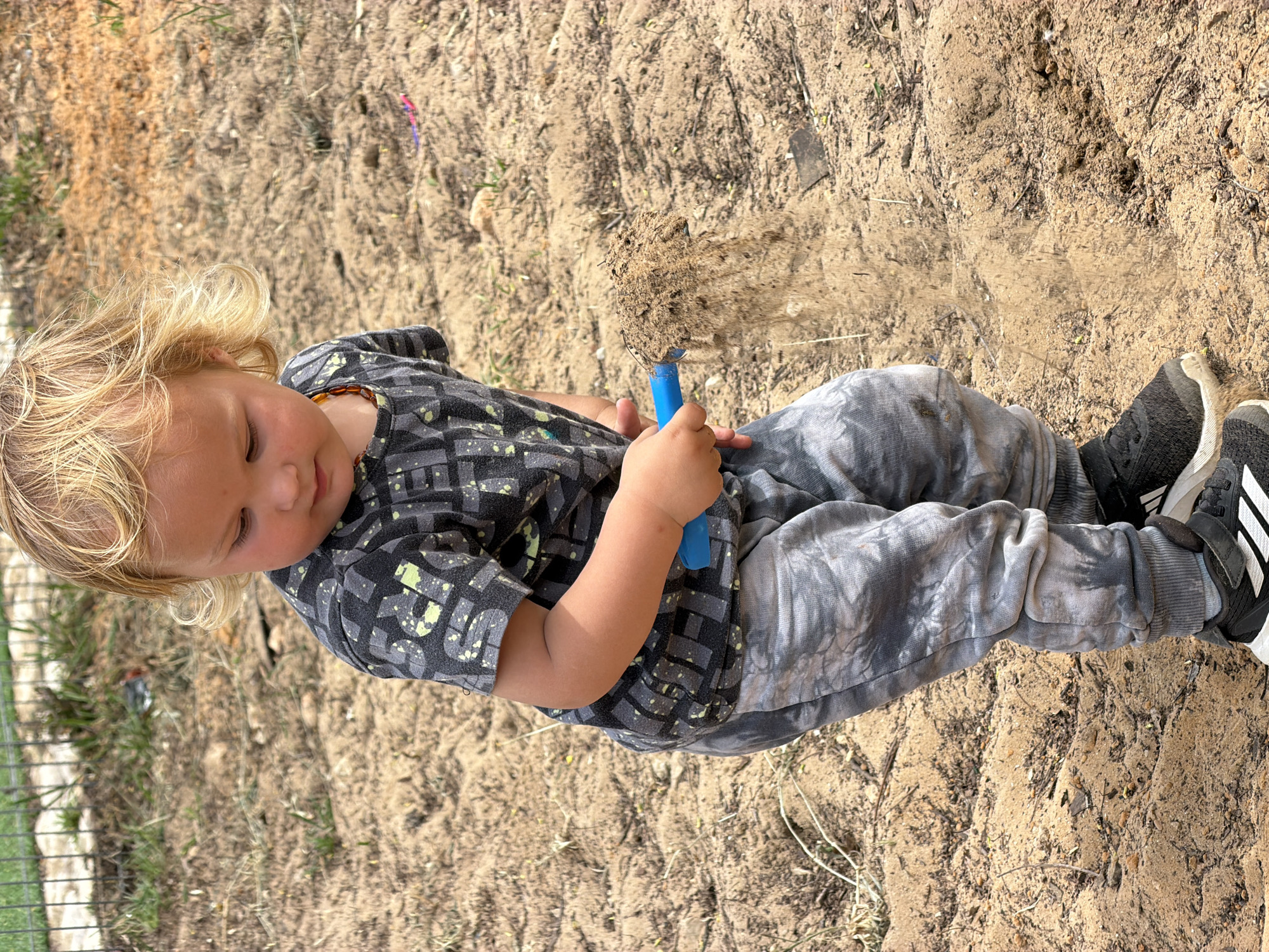 A child with blond hair is playing in the sand at what appears to be a park or playground in Kiryat Bialik, Israel. The child is holding a blue plastic shovel filled with sand, which is being sifted onto the ground. The child is wearing a patterned short-sleeved shirt, gray tie-dye pants, and black Adidas sneakers. The surrounding environment is sandy and dry.