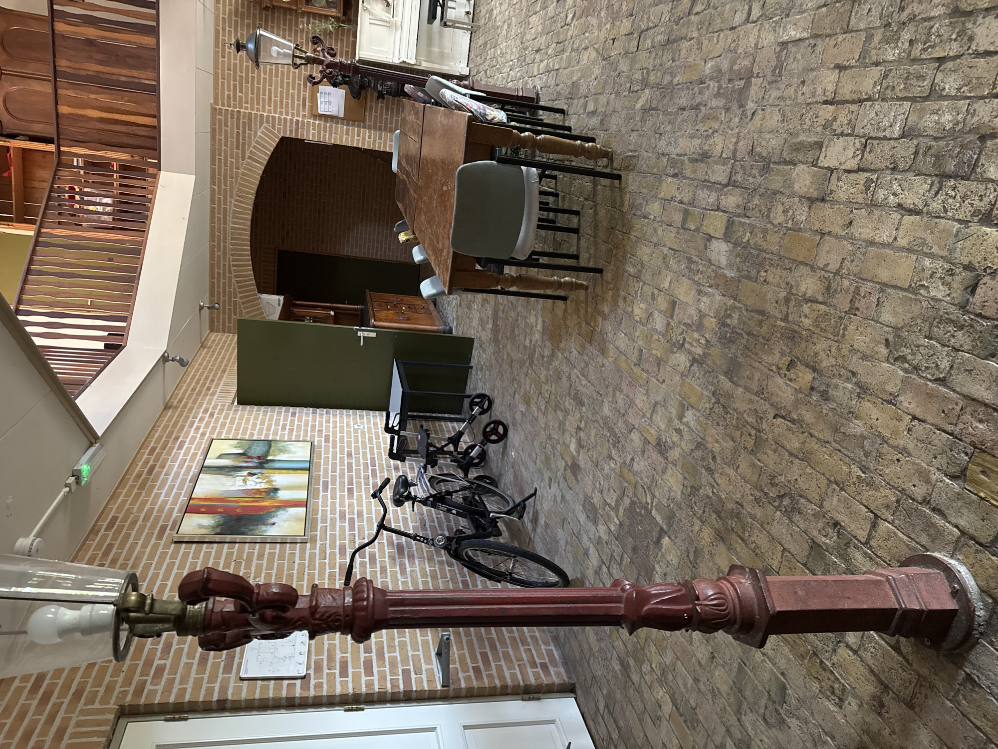 The image shows an indoor scene with brick walls and a brick floor. In the foreground, a red, ornate lamp post stands to the left. To the right of the lamp post, a black bicycle is parked, and behind it is a walker. Further into the scene, a wooden dining table with several chairs is set up. A light-colored piano is visible to the right of the table. The background features an arched doorway and part of a wooden balcony on an upper level. The lighting suggests it is daytime. The location is identified as Pingjum, Netherlands.