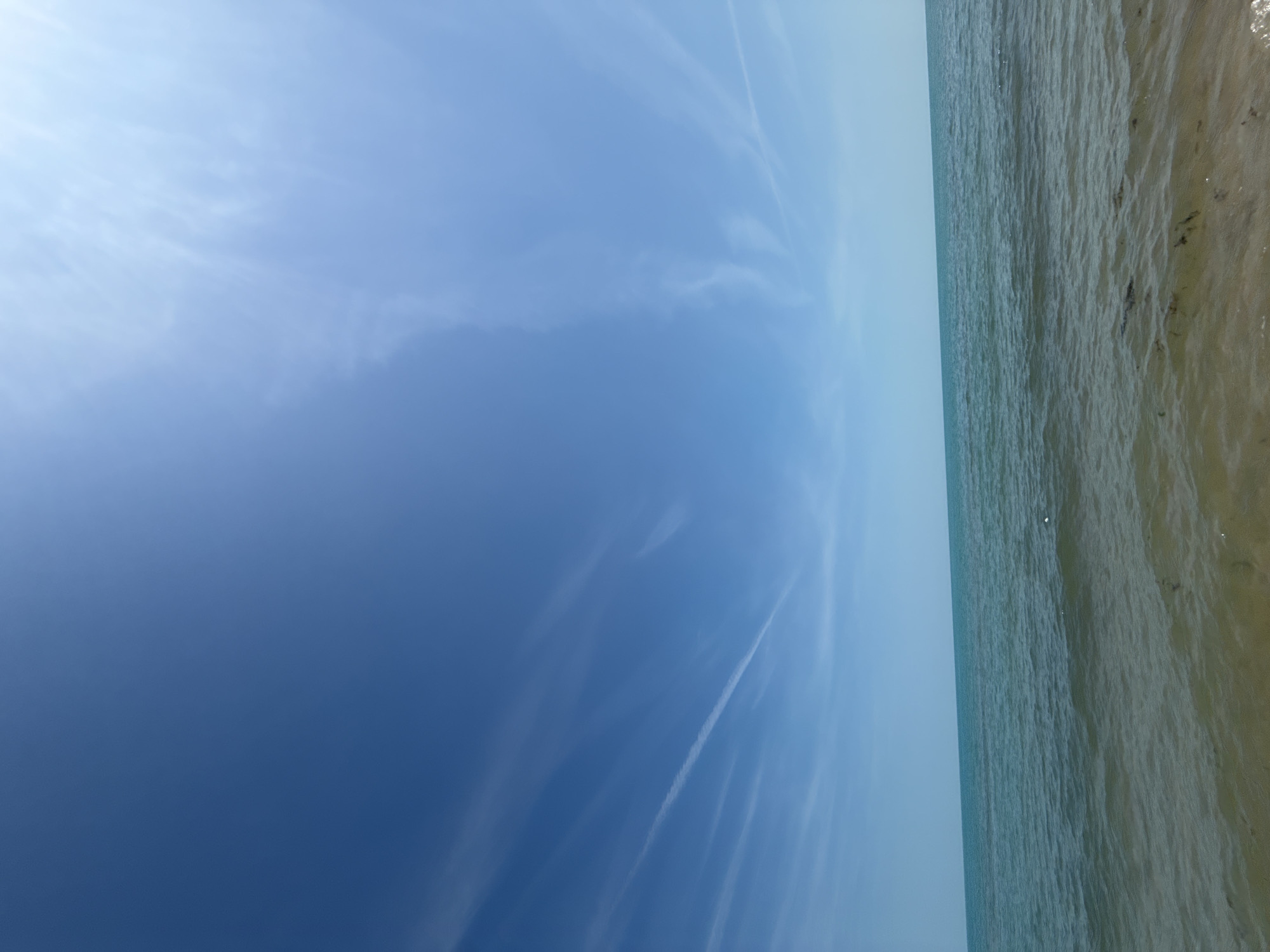 The image captures a clear natural scene in Obrochishte, Bulgaria, focusing on a body of water and the sky above. The upper two-thirds of the frame consist of a bright blue sky, with a gradient from a deeper blue on the left to a lighter, sun-drenched blue on the right. Several faint, wispy white clouds are visible, along with a prominent, straight contrail stretching diagonally across the mid-left portion of the sky. The horizon line is distinct and level. The lower third of the image shows the surface of the sea, exhibiting a range of blues and greens, from a lighter turquoise near the horizon to a darker, more textured green-blue in the foreground. Small ripples and waves indicate gentle water movement. The very bottom of the frame reveals shallow, clear water over a light sandy bottom, with dark patches of what appears to be submerged vegetation or organic debris. No people, animals, or man-made structures are visible.