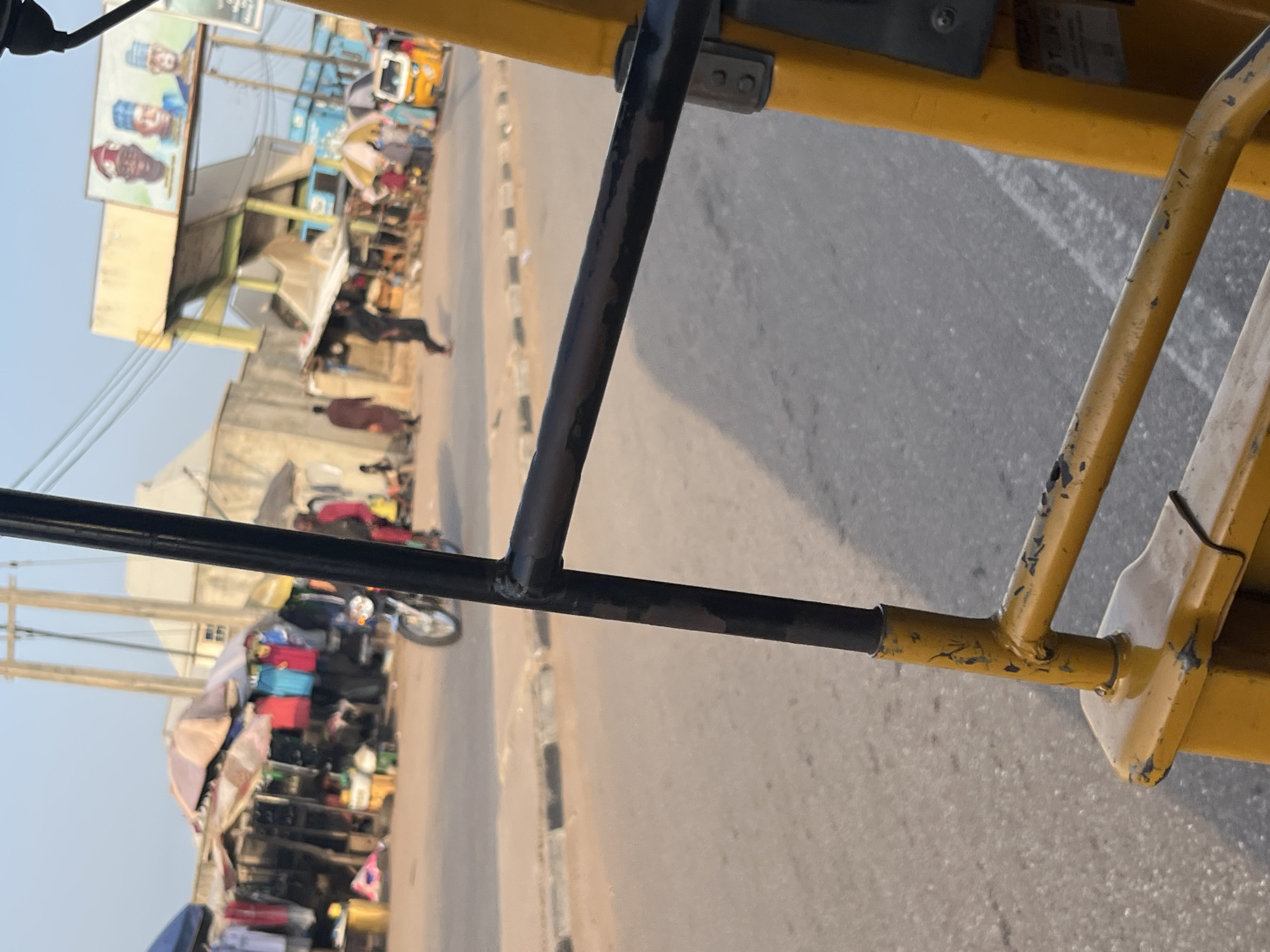 The image captures a street scene from the perspective of a person inside a yellow auto-rickshaw. In the foreground, a section of the auto-rickshaw's yellow frame and a black metal bar are visible. The street is paved and appears to be a road with a curb. In the background, there are market stalls with various items displayed, including clothing. Several people are present, some walking, and one individual is riding a motorcycle. A yellow auto-rickshaw is parked on the side of the road. Above the market stalls, there is a large billboard featuring portraits of three men. The sky is clear and appears to be during daylight. The overall impression is of a bustling marketplace in an urban or semi-urban setting.
