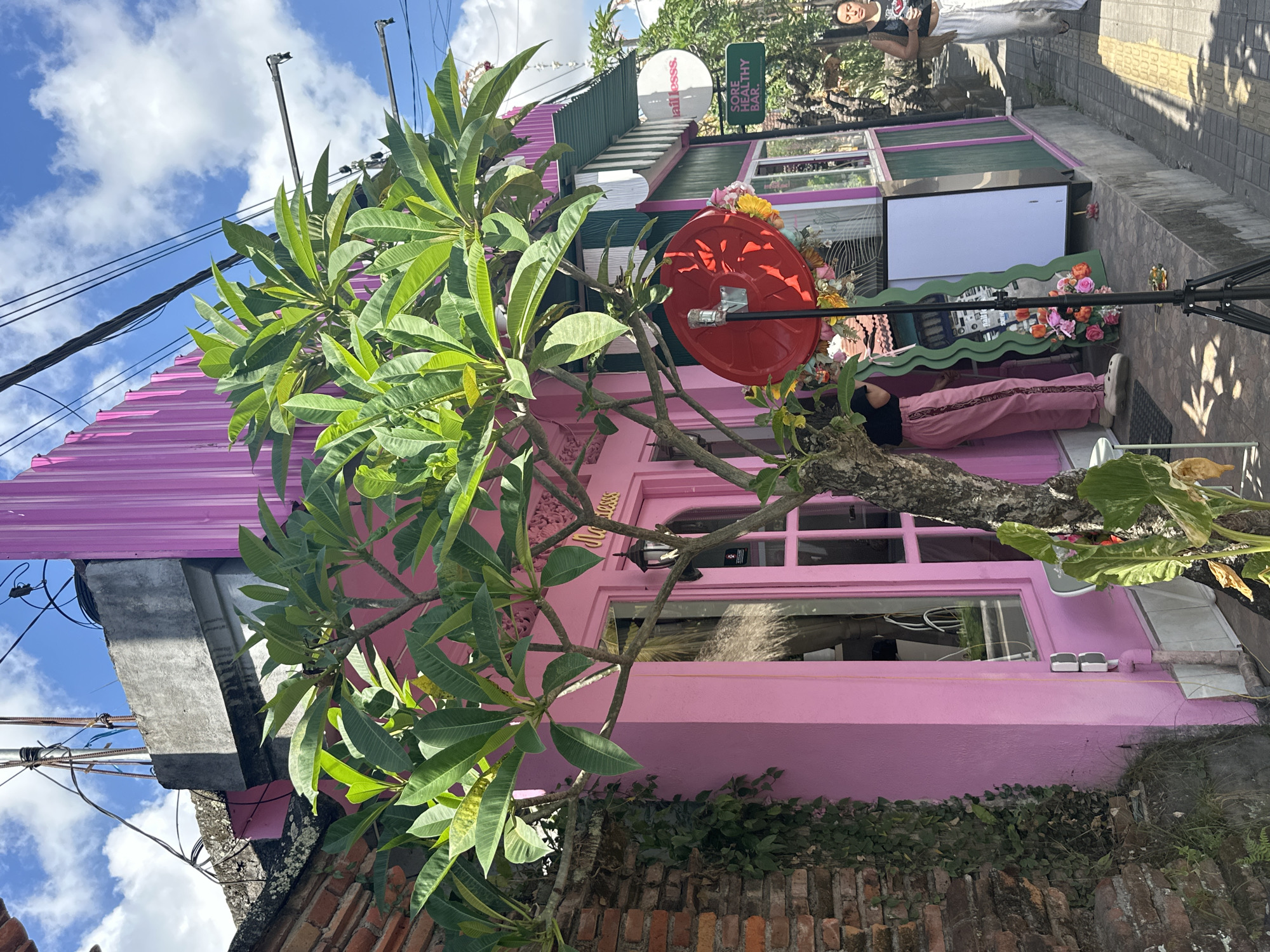 A woman walks down a sunlit sidewalk past a vibrant pink building in Ubud, Indonesia. She wears a black tank top and loose white pants, carrying a bag over her shoulder. The building, adorned with a green awning, has a sign that reads "nailesss" and another below it, "SORE HEALTHY BAR." A tree with lush green leaves partially obscures the building's entrance, which features a pink door and a window. To the left of the building, a brick wall is covered in green ivy. The sky is blue with scattered clouds.